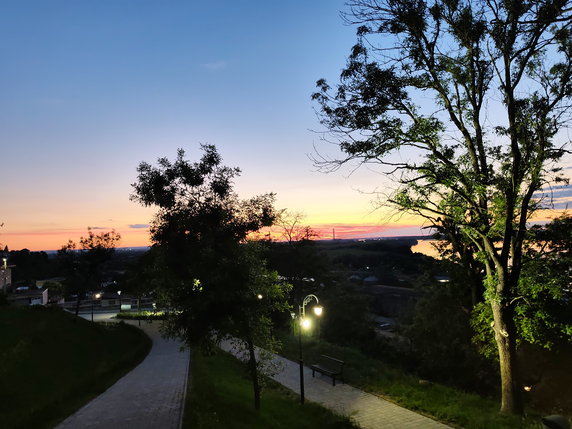 This image depicts a scenic overlook at dusk in the city of Svishtov, Bulgaria. The foreground features a paved walkway winding downhill, bordered by grassy slopes and trees. Several lampposts illuminate the path, casting a warm glow on the surroundings. A park bench is visible near one of the lampposts.
In the middle ground, a small town with residential buildings and more lampposts can be seen nestled amidst the trees and hills. Beyond the town, a river or large body of water reflects the vibrant colors of the sunset. The background is dominated by a vast sky transitioning from a deep blue at the top to hues of orange and pink near the horizon, indicative of twilight. Silhouettes of trees frame the scene, adding depth and a natural element to the urban landscape. The overall atmosphere is peaceful and serene, capturing the beauty of a day's end in Svishtov.