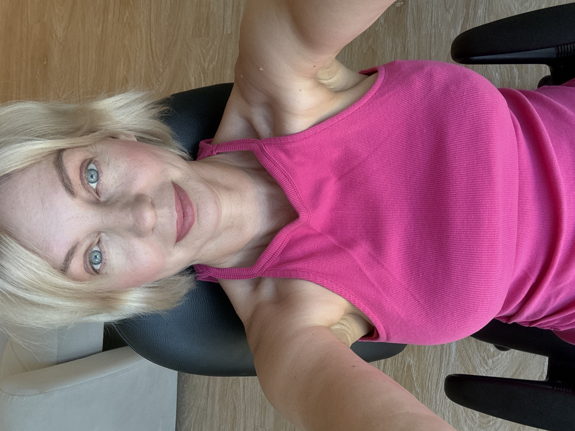 A close-up, overhead selfie shows a blonde woman sitting in a black office chair, wearing a bright pink halter top. Her blue eyes are looking directly at the camera, and she has a slight smile. The background includes a sliver of a white couch on the left and a wooden floor on the right. There is no indication of the city of Sochi, Russia, in this image.