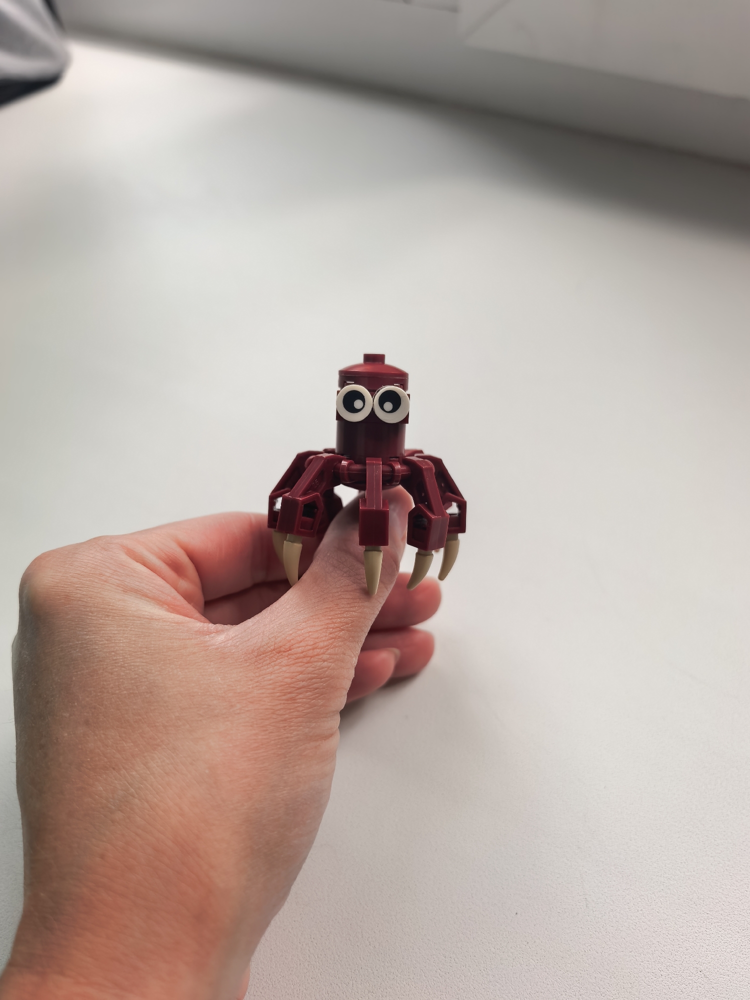 A close-up indoor shot features a person's hand holding a small, red, spider-like construction toy, likely made from building blocks such as LEGO. The toy has a roundish red body with six red clawed legs, each ending in a pointed, light-tan claw. Two large, cartoonish googly eyes are prominently featured on the front of its body. The hand holding the toy is positioned centrally, with the fingers wrapped around the base of the toy. The background consists of a plain, light-colored surface, possibly a white desk or tabletop, under what appears to be bright artificial lighting. A faint shadow falls behind the toy and hand. The upper left corner shows a blurred, darker object or wall element. No text is visible in the image. The setting is an indoor environment, potentially in Sterlitamak, Russia.