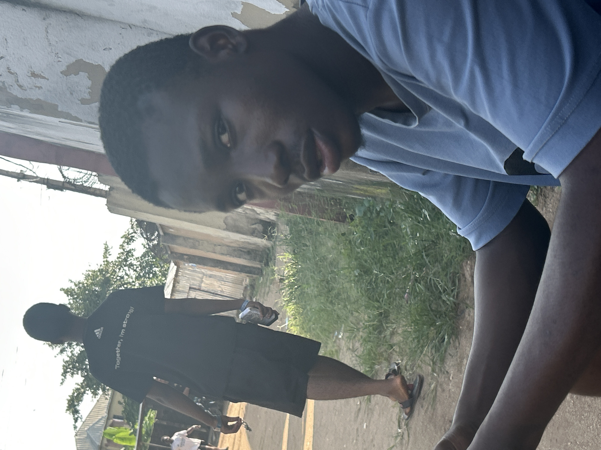 A young Black male in a light blue t-shirt is seated in the foreground, looking towards the camera. Behind him, another Black male wearing a dark t-shirt with the words "Together, I'm strong!" and an Adidas logo is walking away. He is also wearing black shorts and sandals and is holding a white object in his left hand. The background depicts an outdoor scene with a light-colored, weathered wall on the right and some greenery. There are also buildings visible in the distance. The overall setting suggests an urban or semi-urban environment in Ihiagwa, Nigeria.