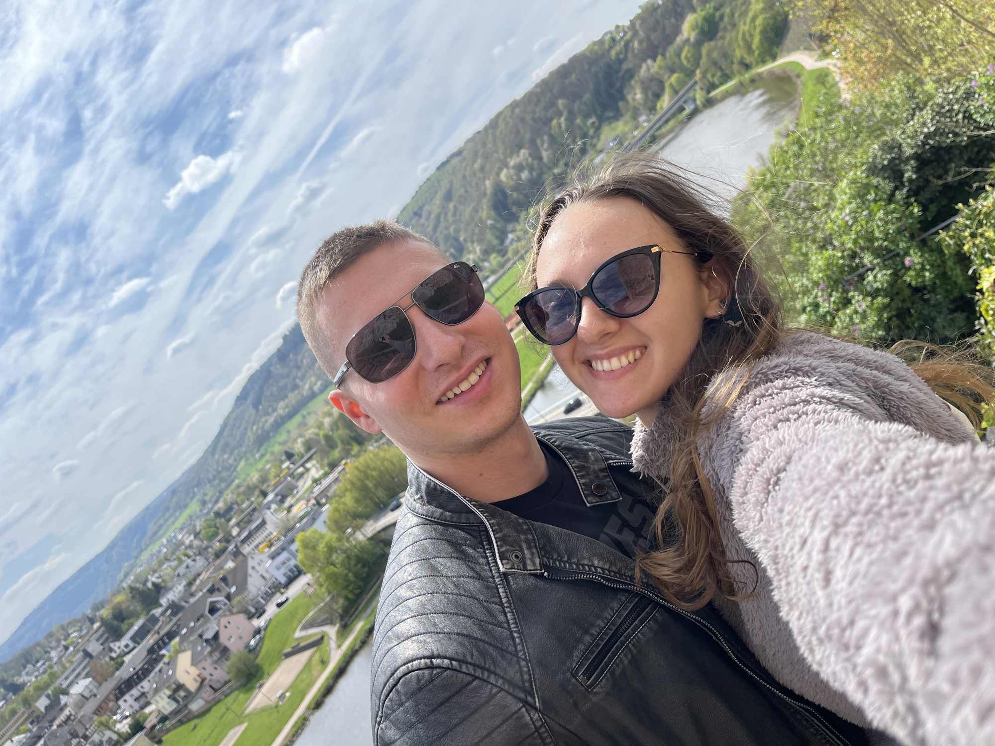 A medium, selfie-style shot shows a man and woman wearing sunglasses smiling, with Saarburg, Germany, visible in the background. The male, on the left, has short brown hair, and wears a black T-shirt under a black leather jacket. The female, on the right, has long, brown hair and wears a grey, fleece jacket. Behind them is a river, lush green vegetation and a portion of the city of Saarburg. A bright sky with scattered clouds is above.