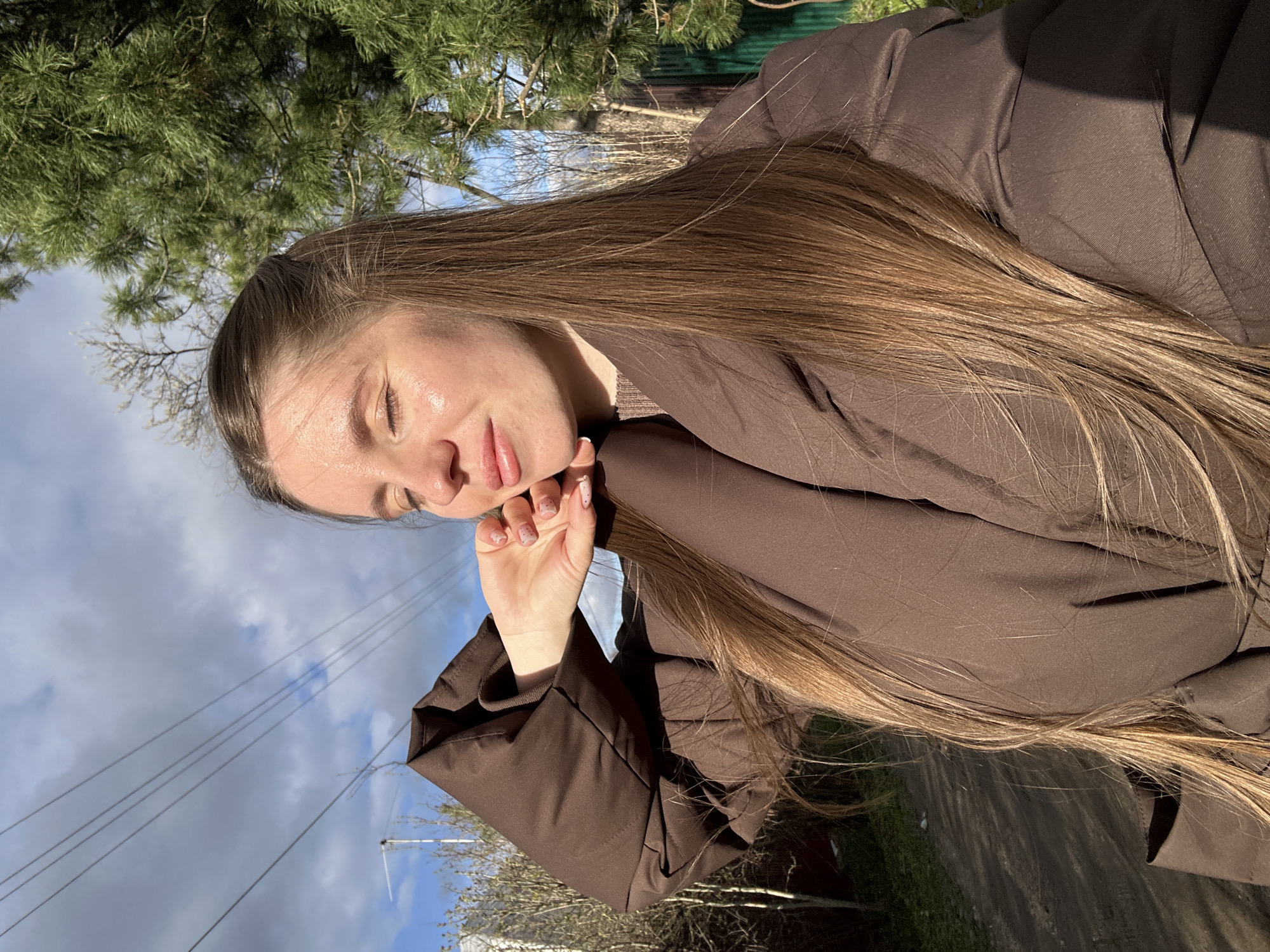 A slightly tilted, outdoor shot captures a young woman from the chest up, positioned against a natural and sky backdrop. She has long, straight brown hair, which flows over her shoulders and down her back. Her eyes are closed or squinted, and her lips are slightly parted, giving her a serene or contemplative expression. Sunlight illuminates her face, highlighting a dewy sheen on her skin, possibly from sun exposure or warmth. She is wearing a brown, seemingly oversized, puffy jacket. One of her hands is raised to her chin/cheek, showcasing nails painted with a light polish and subtle speckles.
The setting appears to be a rural or suburban area in Klin, Russia. In the background, lush green pine trees are visible, along with bare tree branches, suggesting either winter or early spring. The sky is partly cloudy with patches of blue. Power lines crisscross the upper left portion of the image. Faint glimpses of a fence and a dirt path or road can be seen in the distance. The bright sunlight indicates it is daytime, casting clear shadows. There is no visible text in the image.