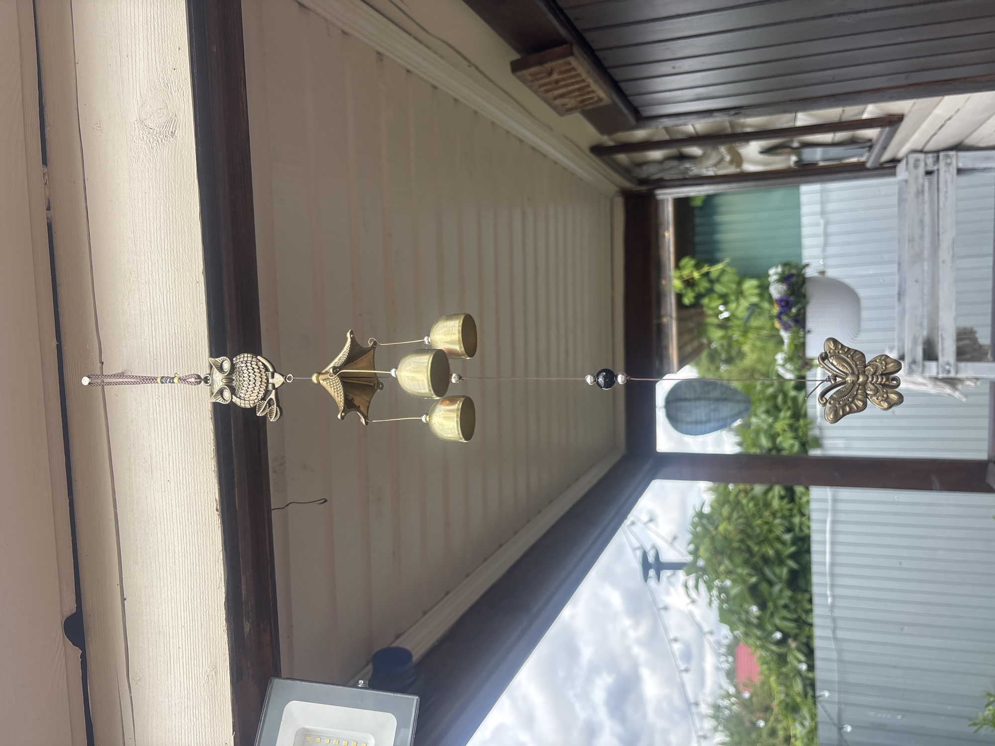 The image captures an outdoor scene under a pale, wood-paneled overhang, likely a porch or veranda. A wind chime, featuring an owl at the top, three brass bells, a black bead, and a butterfly charm at the bottom, hangs from the ceiling. To the left, a modern LED floodlight is attached to the overhang. Beyond the overhang, a garden with lush green trees, a red-roofed structure, and a corrugated metal fence is visible under a cloudy sky. A string of globe lights is strung across the garden area. On the right side of the image, a wooden door with vertical planks is partially visible, with a carved wooden sign that reads "BATHS." A white planter with purple flowers hangs in the background. The overall setting suggests a residential outdoor space in Лотос Снт (марьино), Russia.