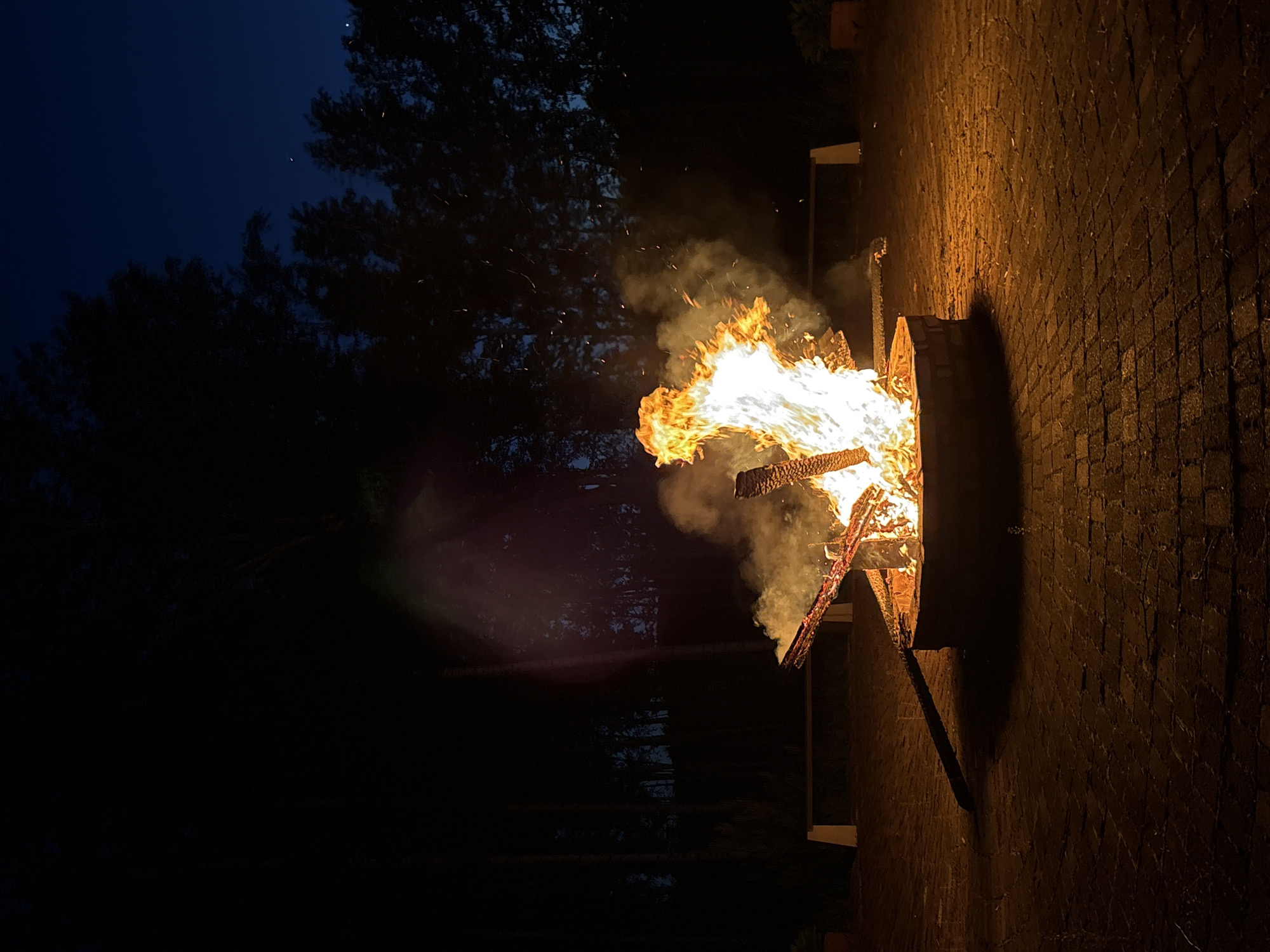 An outdoor brick patio is illuminated by a large bonfire at night. The fire is in a circular brick fire pit, with burning logs and flames reaching upwards. Smoke billows from the fire, and sparks are visible in the air. In the background, silhouetted trees form a dark line against the deep blue night sky. A long, light-colored bench is visible behind the fire pit. The overall impression is one of a quiet, rustic evening.
