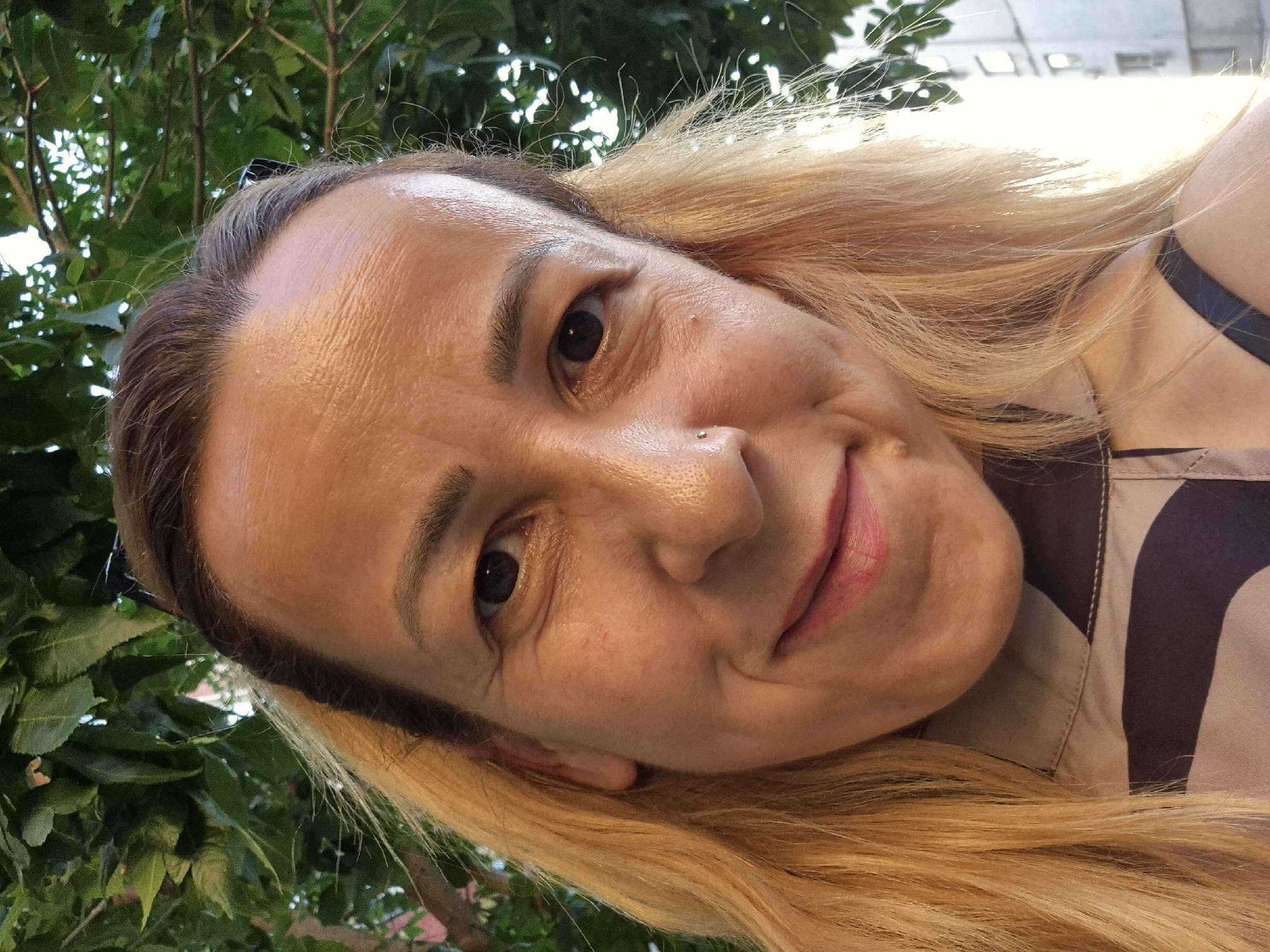A close-up, low-angle portrait captures an adult female with medium skin tone, blonde and light brown hair, and dark eyes, smiling directly at the camera. Her forehead exhibits horizontal lines, and fine lines are visible around her eyes. A small, silver-colored stud piercing is present in her left nostril. Her lips are closed, forming a gentle smile, and show a pink hue. She wears a collared shirt or blouse with a light brown or beige base and abstract, dark brown patterns. A dark strap is visible over her right shoulder. The upper left and top background features dense green foliage, likely tree leaves. The upper right background is brightly lit, revealing the side of a light-colored building with visible windows, indicating an urban or suburban setting. The overall lighting suggests natural outdoor light, with a strong light source originating from the right side of the frame.
