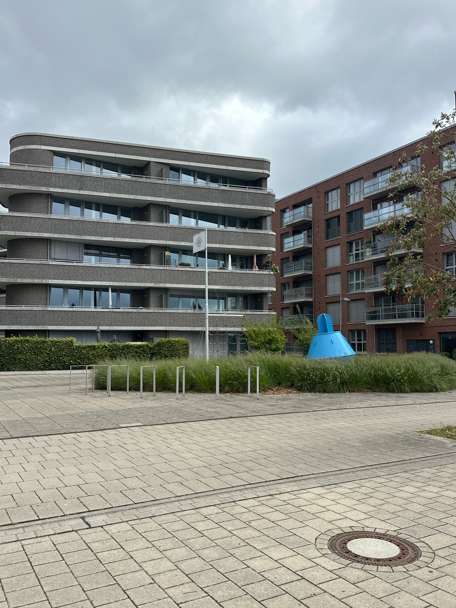The image shows a modern architectural scene in Bremen, Germany. In the foreground, there is a paved walkway with a manhole cover. Behind the walkway, there is a landscaped area with ornamental grass and several thin, vertical metal poles. Further back, two apartment buildings are visible. The building on the left is made of dark brick with curved balconies and rows of windows. The building on the right is made of red brick and has balconies with glass railings. In the landscaped area between the buildings, there is a large, bright blue, conical sculpture. The sky is overcast.