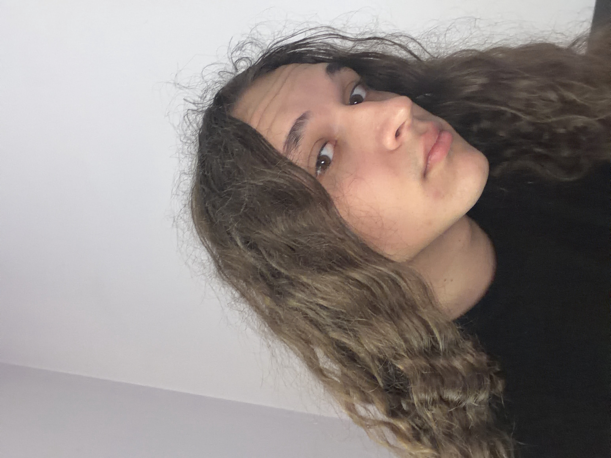 The image is a close-up of a person with long, curly brown hair. The person is facing slightly to the side, with their face visible from the nose to the forehead. The background appears to be a plain white wall. The person is wearing a black shirt. There are no visible landmarks or other details to suggest a location in Asenovgrad, Bulgaria.