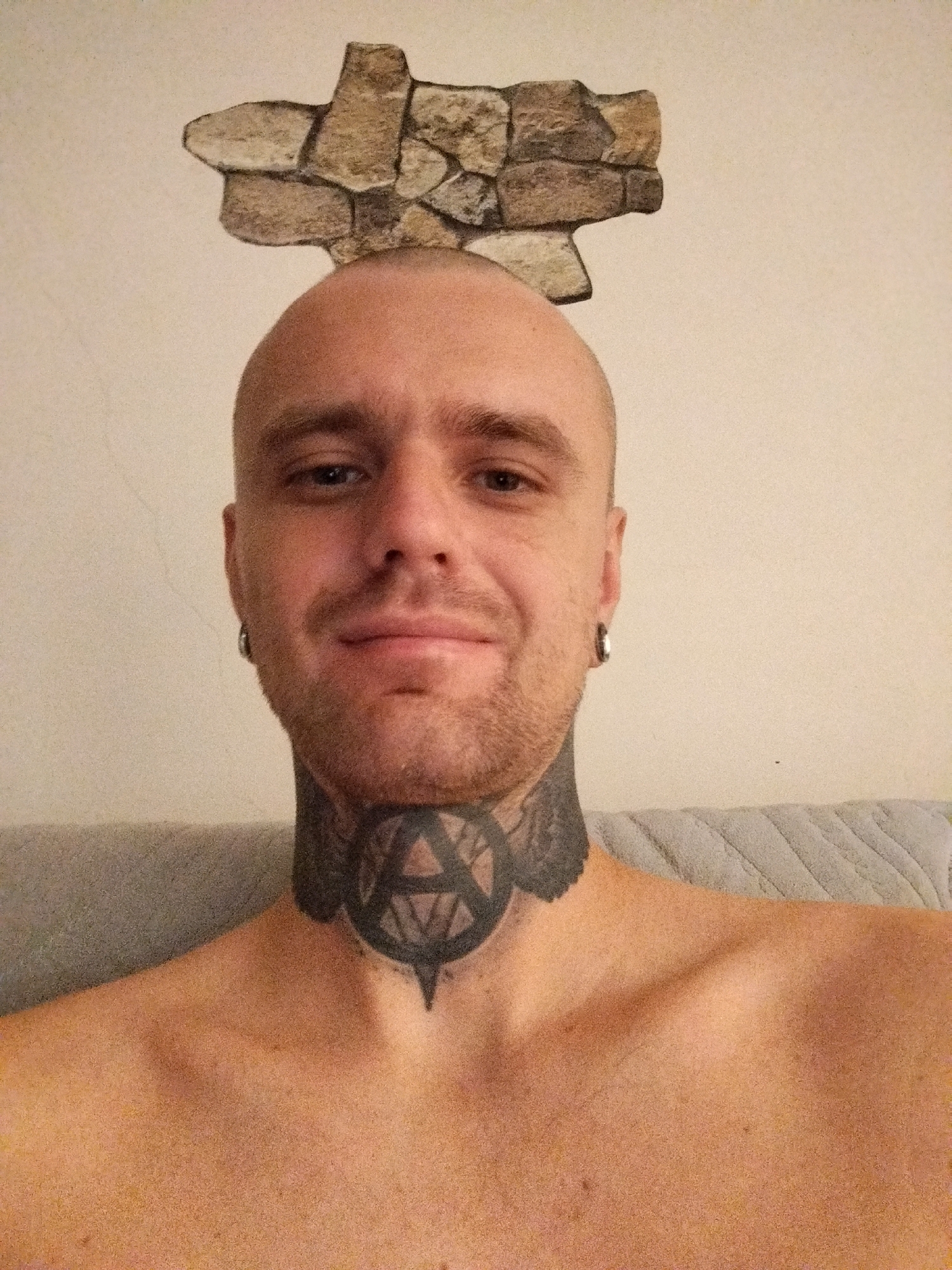 A close-up, frontal, waist-up selfie shows a bald, fair-skinned man with a light beard and a tattoo around his neck and upper chest. He is wearing a silver ear gauge in his left ear. He is shirtless. In the background, there is a light-colored wall with a decorative element resembling rough stones at the top, and a textured, light gray surface, possibly a sofa or chair, occupying the lower part of the frame. The man's eyes are open, and he is looking directly at the camera with a neutral expression. The lighting appears to be natural, coming from the front. The setting is indoors, likely a residential or personal space, and there is no indication of the city of Batumi, Georgia, or any urban elements.