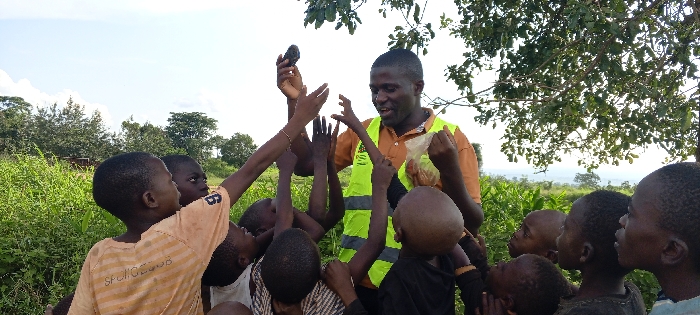 The image depicts an outdoor scene in Ngalula, Democratic Republic of the Congo. Centrally positioned, a dark-skinned adult male wearing an orange shirt and a bright yellow-green reflective vest interacts with approximately seven to nine dark-skinned children of various ages, appearing to be under 12 years old. The adult male holds a dark, rectangular object, consistent with a mobile phone, in his raised right hand and a clear plastic bag containing multiple small, brownish items in his left hand. His mouth is open, suggesting speech or laughter. Several children have their arms raised and hands extended towards the adult male. The setting features dense green vegetation in the foreground and midground, with trees visible in the background against a bright sky and a hazy distant horizon.