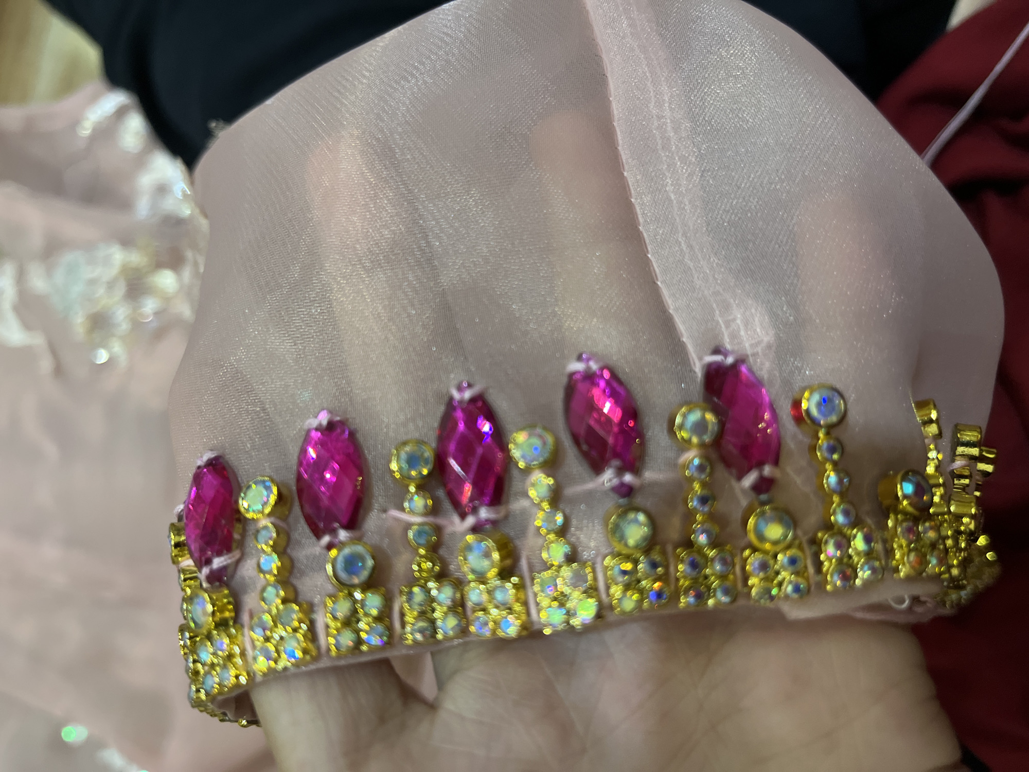 The image displays a close-up view of a pink, sheer fabric adorned with a decorative trim. The trim features a repeating pattern of gold settings encasing small, iridescent circular stones, interspersed with larger, faceted fuchsia-colored gemstones. The fabric is being held by a person's hand, partially visible in the lower left corner. The background is blurred, showing hints of other fabric textures and colors, possibly indicating a tailoring or craft setting. The lighting suggests an indoor environment. The image does not contain any information about the city of Palopo, Indonesia.