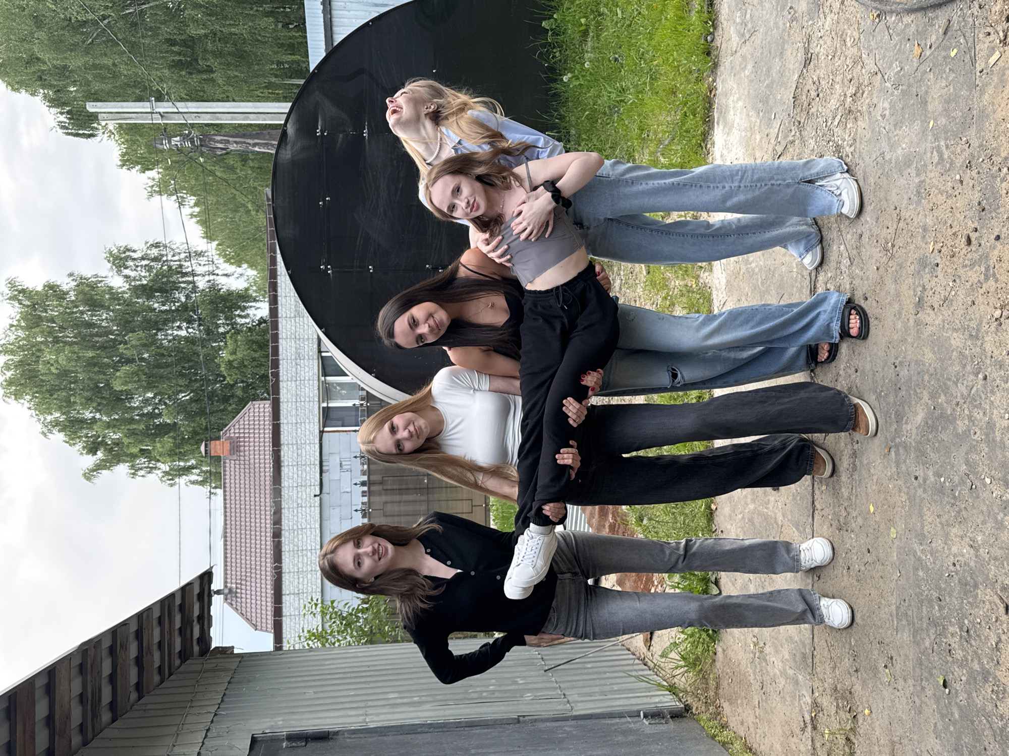Four young women are posing for a photo outdoors. The woman on the far left is standing with her hands on her hips, looking at the camera. The second woman from the left is standing, holding the legs of a fifth person, who is lying horizontally across the laps of the other two women on the right. The woman on the far right is leaning back, laughing. The scene appears to be in a backyard with a building on the left and a black, arched structure in the background. The ground is concrete and dirt, with grass to the right. The weather appears overcast.