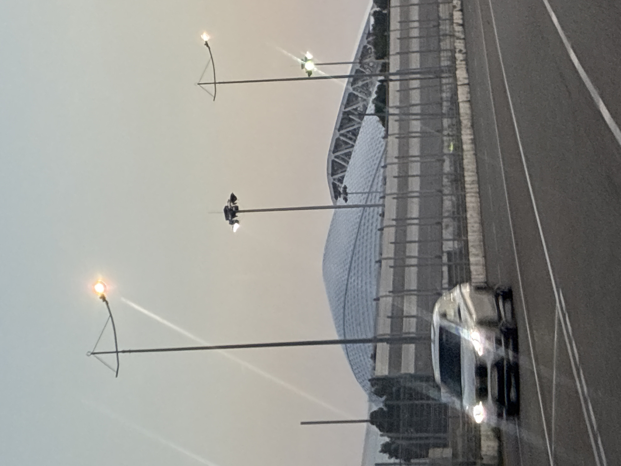 The image captures a view of the Fisht Olympic Stadium in Sirius, Russia, during what appears to be either dawn or dusk, given the soft, hazy lighting. In the foreground, a white car is driving on a road with visible lane markings. The car's headlights are illuminated. Behind the road, a grey, slatted fence runs horizontally across the frame. Beyond the fence, the distinctive white, dome-like structure of the Fisht Stadium is prominent, with its arched roof structure visible towards the right side of the stadium. Several tall light poles, some with illuminated lamps, are scattered throughout the scene, with one in the left mid-ground and two in the right mid-ground. The sky is a pale, gradient color, suggesting either early morning or late evening light. Some faint diagonal lines are visible in the sky, possibly reflections or lens flares.