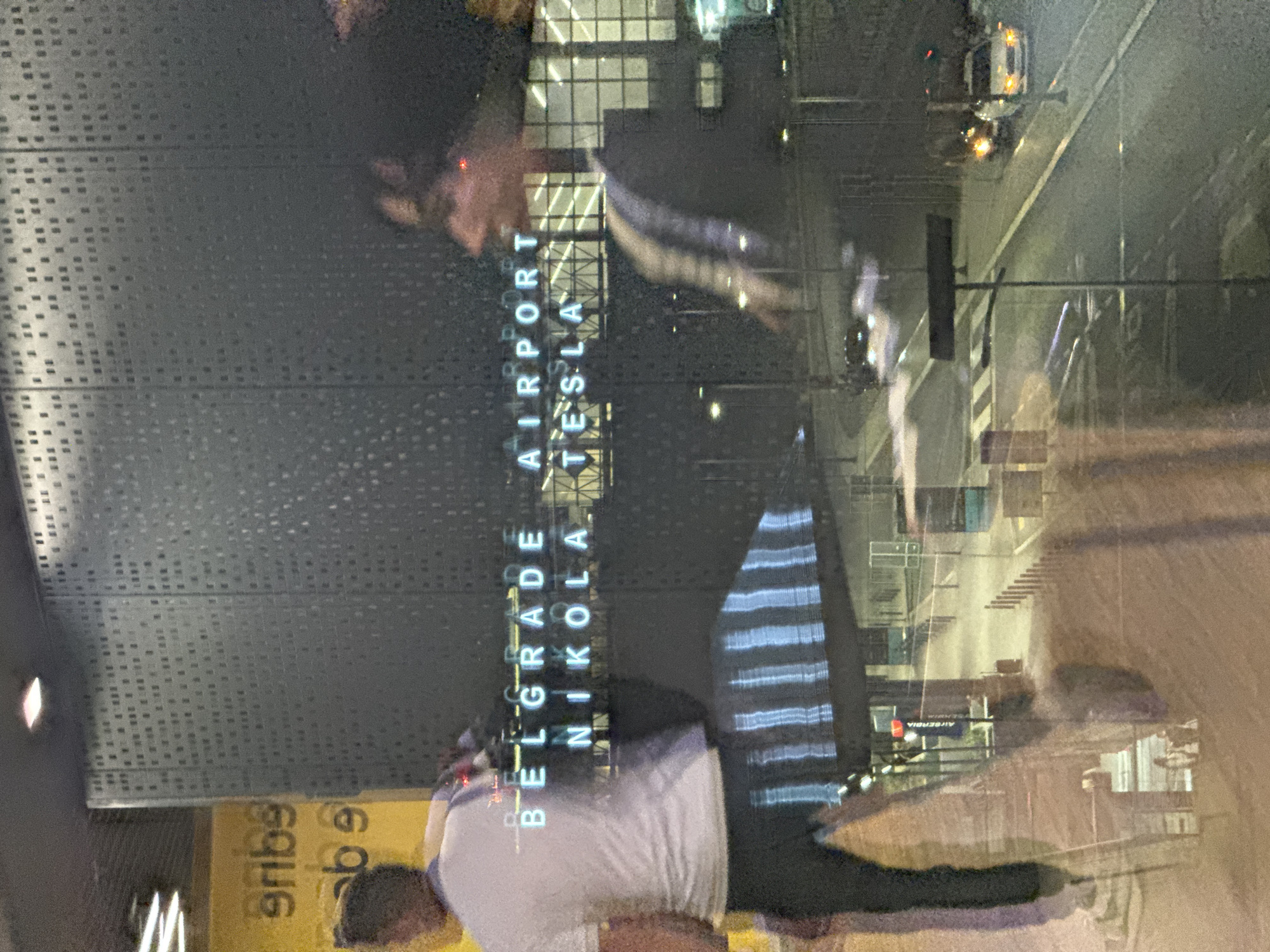 The image shows a view from inside Belgrade Nikola Tesla Airport (BEG) in Surcin Urban Municipality, Serbia. The reflection in the window shows a person looking down, wearing a white t-shirt and dark pants, and another person wearing a dark jacket with light stripes. The airport's exterior is visible in the reflection, including the "BELGRADE AIRPORT NIKOLA TESLA" sign in vertical white illuminated letters. There is a roadway with vehicles, including a bright white vehicle, and various buildings visible through the window. A yellow sign with black writing, partially cut off, is visible in the lower left of the image. The window itself has a pattern of small square perforations.