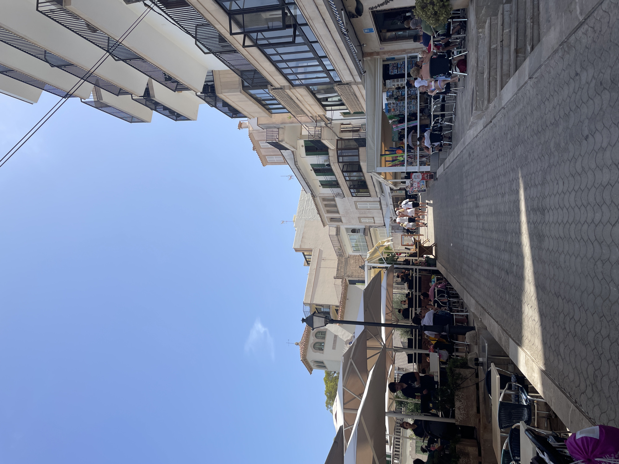 The image captures a sunlit street scene in Santanyí, Spain, likely within the port area given the architecture and ambiance. The perspective is from a pedestrian walkway, looking uphill towards a cluster of buildings and a bright blue sky.
On the right side of the street, a modern, multi-story building dominates the view, featuring multiple balconies with black railings. Its facade is a light color, with large windows. Below this building, several businesses appear to be operating, with awnings extended over seating areas. People are seated at tables outside these establishments, some dining and others engaged in conversation. Waitstaff are visible, attending to customers.
On the left side of the street, a row of older, more traditional buildings is present. These structures have a lighter, textured facade and feature balconies, some with wrought-iron railings. Similar to the right side, there are outdoor seating areas for cafes or restaurants, with umbrellas providing shade. People are also seated at these tables, and a few individuals are walking along the path.
The walkway itself is paved with interlocking, light-colored stones, arranged in a curved pattern. The ground slopes upwards. A street lamp is visible in the middle ground. The overall atmosphere is one of a vibrant, bustling European street, likely during daytime hours, with clear weather.
