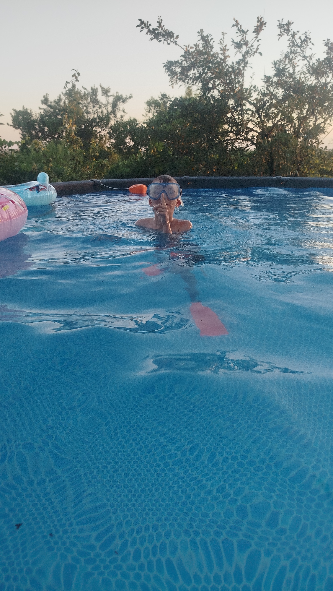 A person wearing swimming goggles and flippers is in a swimming pool. The person is partially submerged in the water and has their finger to their lips, as if signaling for quiet. There are inflatable toys floating in the pool, including a pink one on the left and a blue one behind the person. The water is blue and reflects the sunlight. In the background, there are trees and a clear sky. The image was taken in Kostiantynivka, Ukraine.
