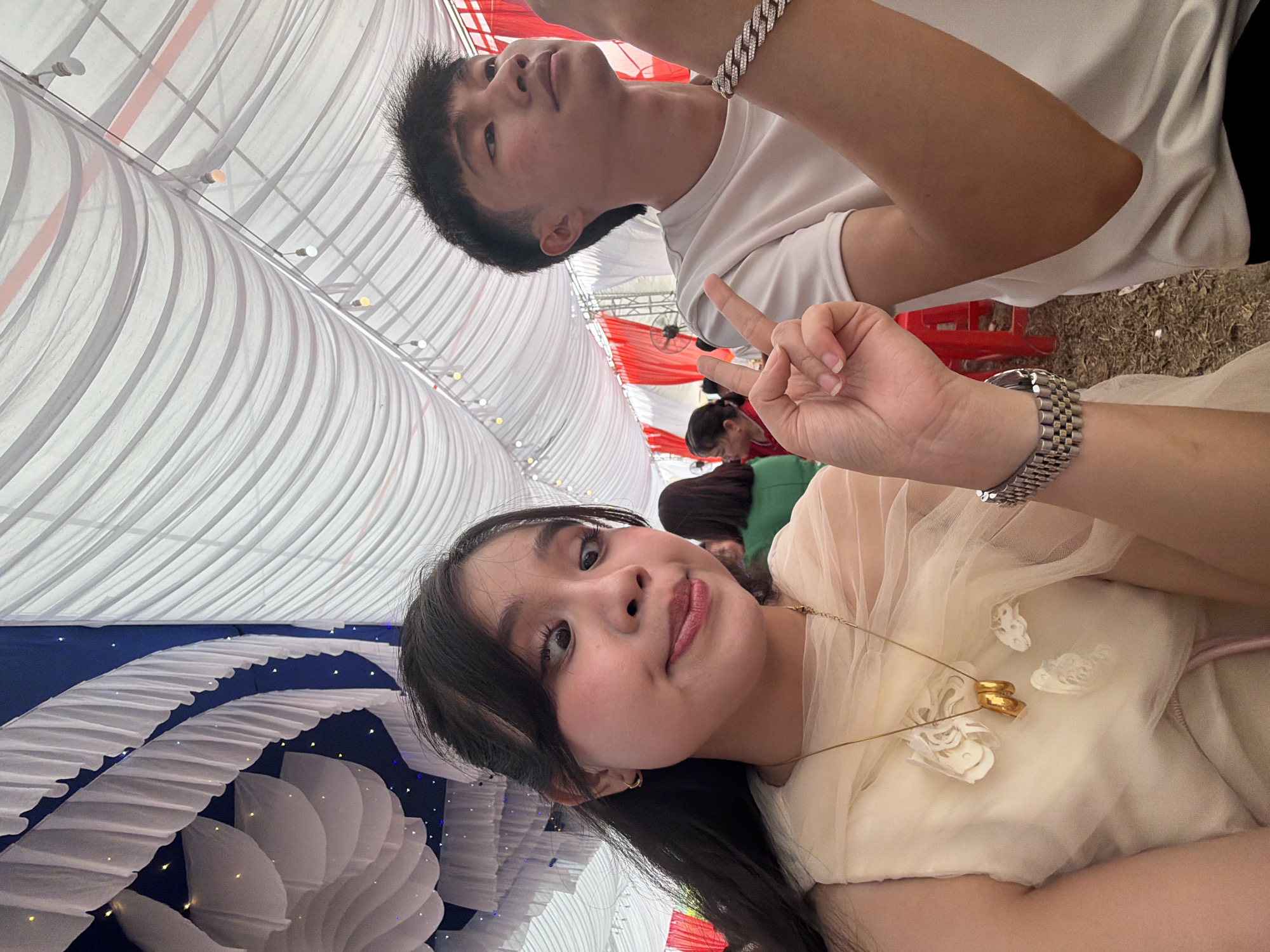 This image captures two young individuals posing at an indoor or covered outdoor event, likely in Tay Son Commune, Vietnam.
In the foreground, a young woman with long dark hair is looking directly at the camera, wearing a light-colored, possibly cream or light yellow, draped garment. She has a subtle smile or pout and holds her left hand up, forming a peace sign. Her left wrist displays a silver-colored watch with a link bracelet, and around her neck, she wears a delicate gold chain necklace with two gold rings as a pendant, resting on her chest.
Behind her and slightly above, a young man with short dark hair is visible, wearing a plain white t-shirt. He has a silver-colored link bracelet on his left wrist and is looking upwards and to his left with a neutral expression.
The setting appears to be a decorated venue, possibly a large tent or event hall. The ceiling is draped with white fabric, creating a soft, pleated effect, and small lights are interspersed within the drapes. In the background, there are sections of blue fabric decorated with white ruffled patterns and more small lights, as well as red and white fabric decorations further back. Other indistinct figures are visible in the distance, suggesting a gathering or celebration. The lighting indicates it is daytime, with bright ambient light illuminating the space. No visible text is present.