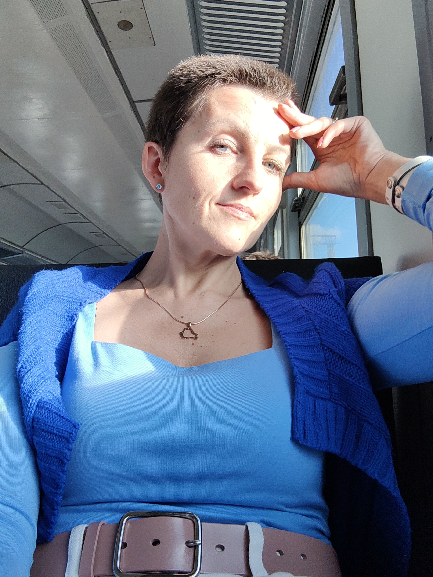 A light-skinned woman with a short haircut is captured in a selfie taken inside a train carriage, likely in Minsk, Belarus. She is wearing a bright blue long-sleeved top and a blue knitted cardigan draped over her shoulders. A rose-colored belt with a silver buckle cinches her waist. Her right hand is resting on her forehead, and a silver bracelet is visible on her wrist. A thin gold chain with a triangular pendant hangs around her neck. A small blue earring adorns her left ear. The background shows the interior of the train, with its ceiling and a window revealing a bright sky outside. The lighting suggests it is daytime, with sunlight casting shadows on her face.