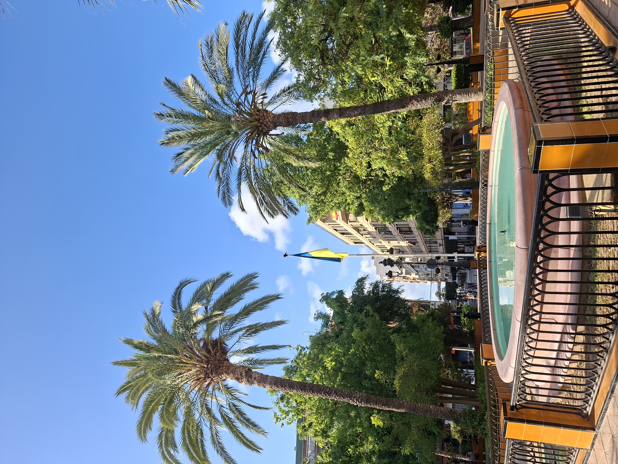 The image depicts a sunny day in Torrevieja, Spain. In the foreground, a circular fountain with turquoise water is surrounded by a black wrought-iron fence with ornate scrollwork and topped with orange tiled posts. Beyond the fountain, lush green trees and palm trees create a vibrant urban park setting. In the background, buildings line a street, and a Ukrainian flag is prominently displayed on a flagpole, fluttering in the gentle breeze. The sky is a brilliant blue with scattered white clouds. A few people are visible in the distance, walking along the street. The overall impression is one of a peaceful and lively city square.