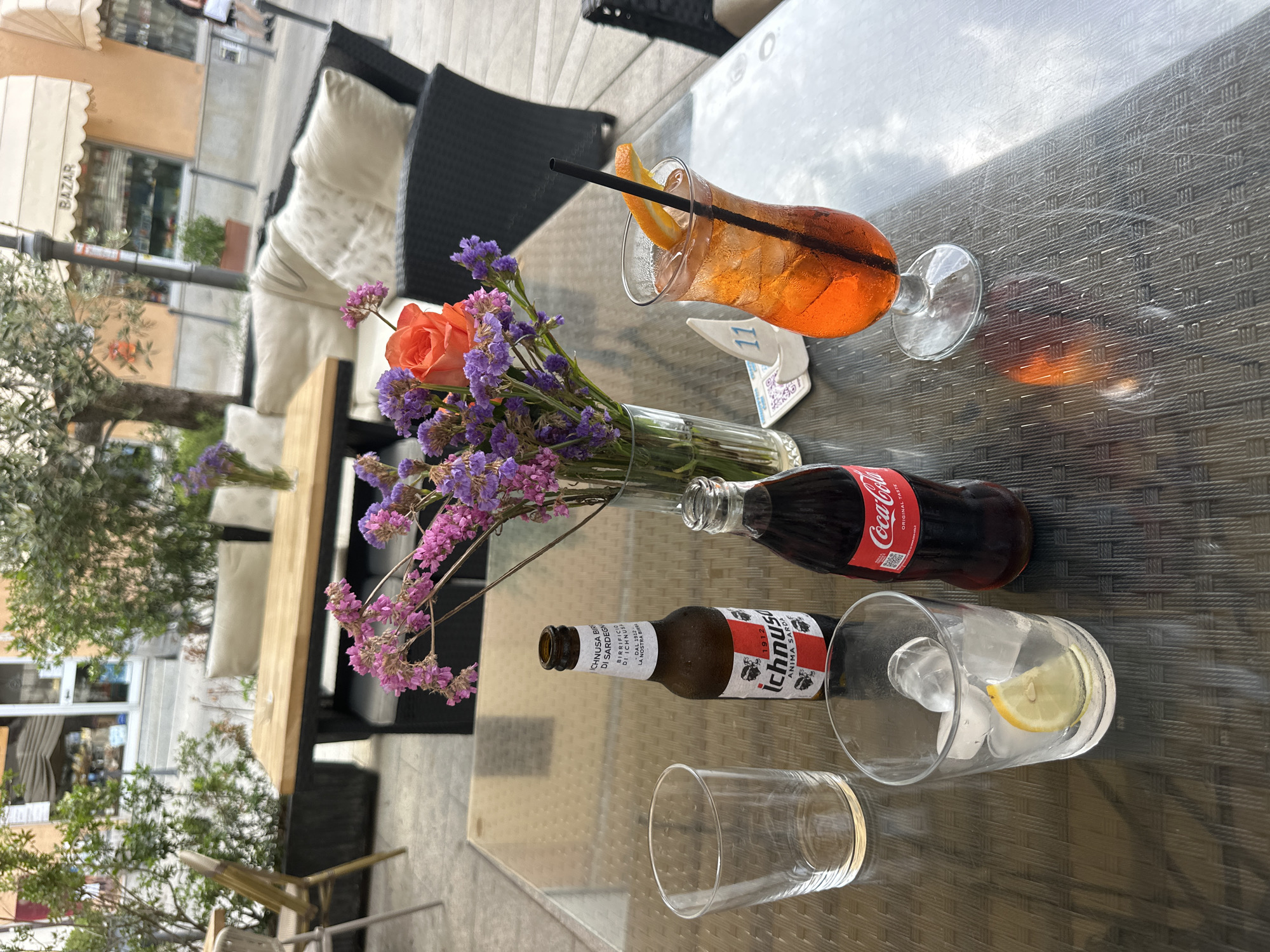 An outdoor table at a cafe in Villasimius, Italy, is set with drinks and a floral arrangement. The table surface is textured and reflective, with the dappled light of an overcast day. On the table, a bottle of Ichnusa Birra di Sardegna beer, a bottle of Coca-Cola, and a glass filled with ice and a lemon slice are visible. A vibrantly colored orange cocktail with a straw and an orange slice garnish is also present. A vase of purple and pink dried flowers, with a single orange rose, adds a decorative element. In the background, outdoor seating with cushions and potted plants are seen, along with a storefront with awnings and the word "BAZAR" visible. A woman in dark clothing and a white top is walking in the distance.