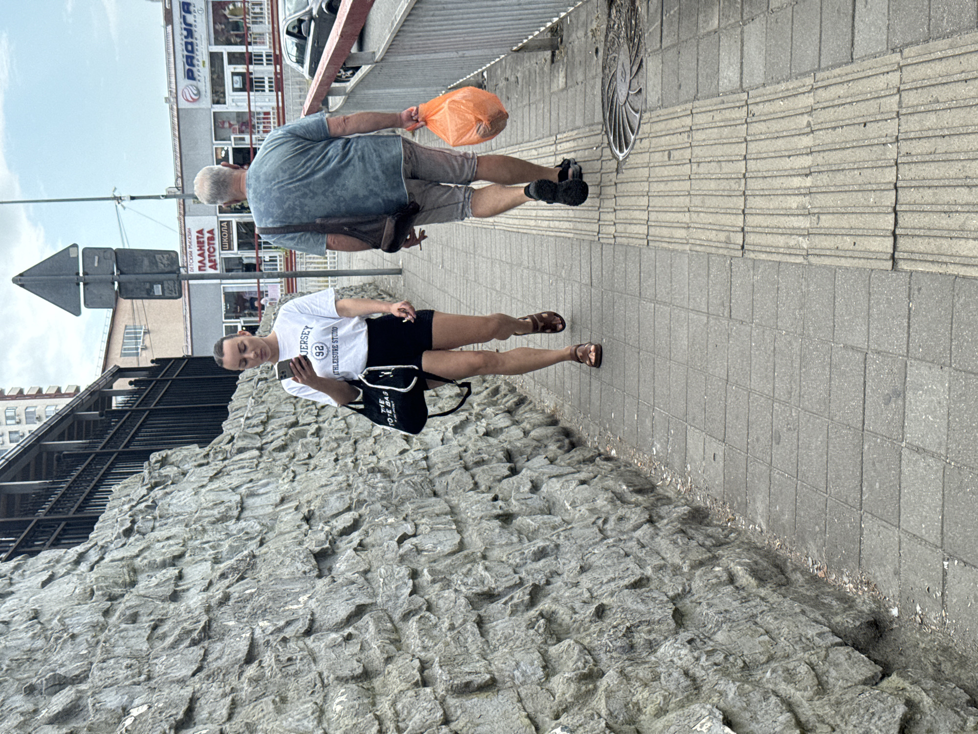 This media file captures two individuals in an urban outdoor setting, likely in Sochi, Russia, given the provided context. The scene appears to be a sidewalk adjacent to a building and a textured stone wall.
In the foreground, a person is shown from the waist down, wearing a white t-shirt with the text "JERSEY 92 ATHLEISURE STUDIO" visible. They are also wearing black shorts and brown sandals, and are carrying a black bag with white lettering that includes "TO THE TOTE BAG". This individual is holding a smartphone and appears to be standing or walking on the sidewalk.
Behind them and slightly further away, another person is walking, seen from the waist up. This individual is wearing a blue t-shirt and grey shorts and is carrying an orange plastic bag in their right hand. A brown bag strap is visible across their shoulder. This person is also walking on the sidewalk, near a traffic sign.
The setting suggests a daytime scene under an overcast sky, as indicated by the lighting and the ambient brightness. The background shows buildings with visible signage, including "РАДУГА Курортные товары" (Raduga Resort Goods) and "ДЕТСКИЙ МАГАЗИН ПЛАНЕТА ДЕТСТВА ШКОЛА" (Children's Store Planet of Childhood School). The presence of these signs and the style of the buildings strongly points to a Russian location. The sidewalk is made of concrete tiles, and there's a distinct textured stone wall to the left of the frame. The overall atmosphere is casual and everyday, with no indication of a specific event beyond people going about their day.