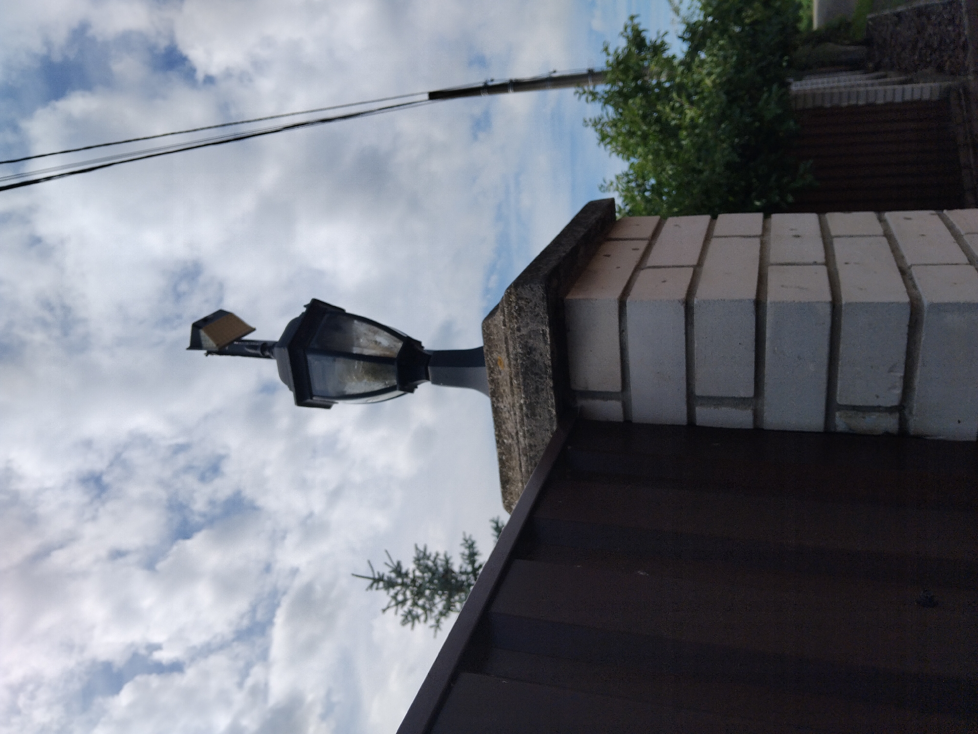 This media file depicts an outdoor scene, likely a residential area in Kyiv, Ukraine. The primary subjects are a fence, a lamppost, and the sky. The fence appears to be constructed from dark brown material, possibly wood or metal, and is topped with a section of light-colored brickwork, suggesting a boundary or entrance to a property. A classic-style lamppost, with a black metal frame and glass panes, is mounted on a concrete base, protruding from the fence. Above the fence and lamppost, a cloudy sky dominates the frame. The clouds are predominantly white and billowy, interspersed with patches of blue sky, indicating a partly cloudy day. A few trees with green foliage are visible in the background, peeking over the fence and suggesting a relatively lush environment. Power lines also traverse the sky, a common sight in urban or suburban landscapes. The image is taken from a low angle, looking upwards towards the sky, which emphasizes the height of the fence and lamppost and the expanse of the sky. There is no visible text in the image, nor any discernible activity or event taking place. The overall impression is one of a calm, overcast day.