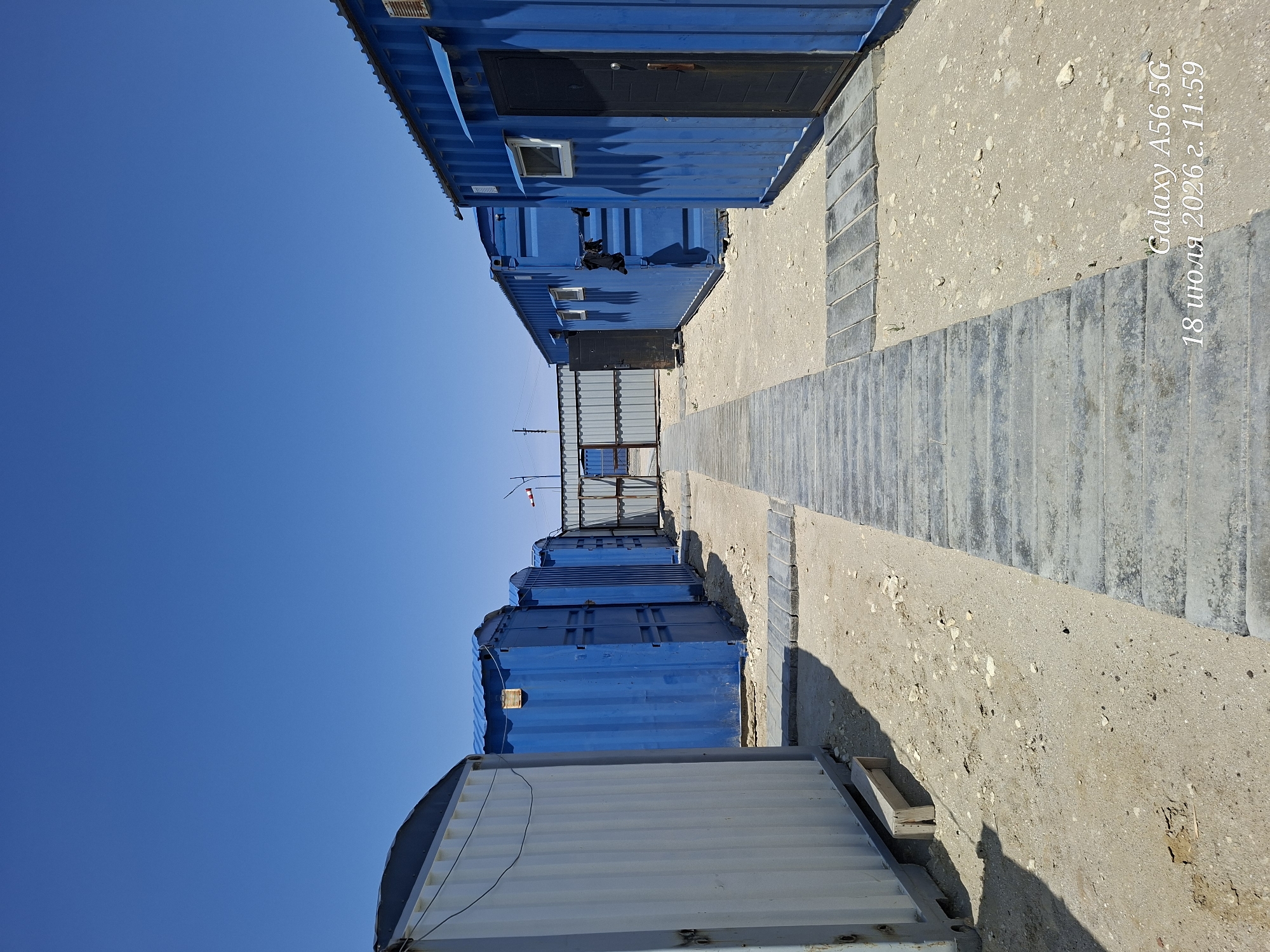 A wide-angle, eye-level exterior shot captures a row of blue shipping container-like structures aligned parallel to a walkway made of concrete slabs. The structures appear to be temporary modular buildings, possibly housing or offices, as indicated by the windows and doors visible on some. The walkway leads towards a chain-link fence in the distance, beyond which is a barren, sandy landscape. The sky above is a clear, bright blue. The image appears to be taken on a sunny day, with shadows cast by the buildings. A timestamp at the bottom right of the image reads "Galaxy A56 5G 18 июля 2026 г. 11:59", indicating the date and time the photo was taken. The environment suggests a remote or construction site location, possibly in a desert or semi-arid region. There are no people visible in the image.