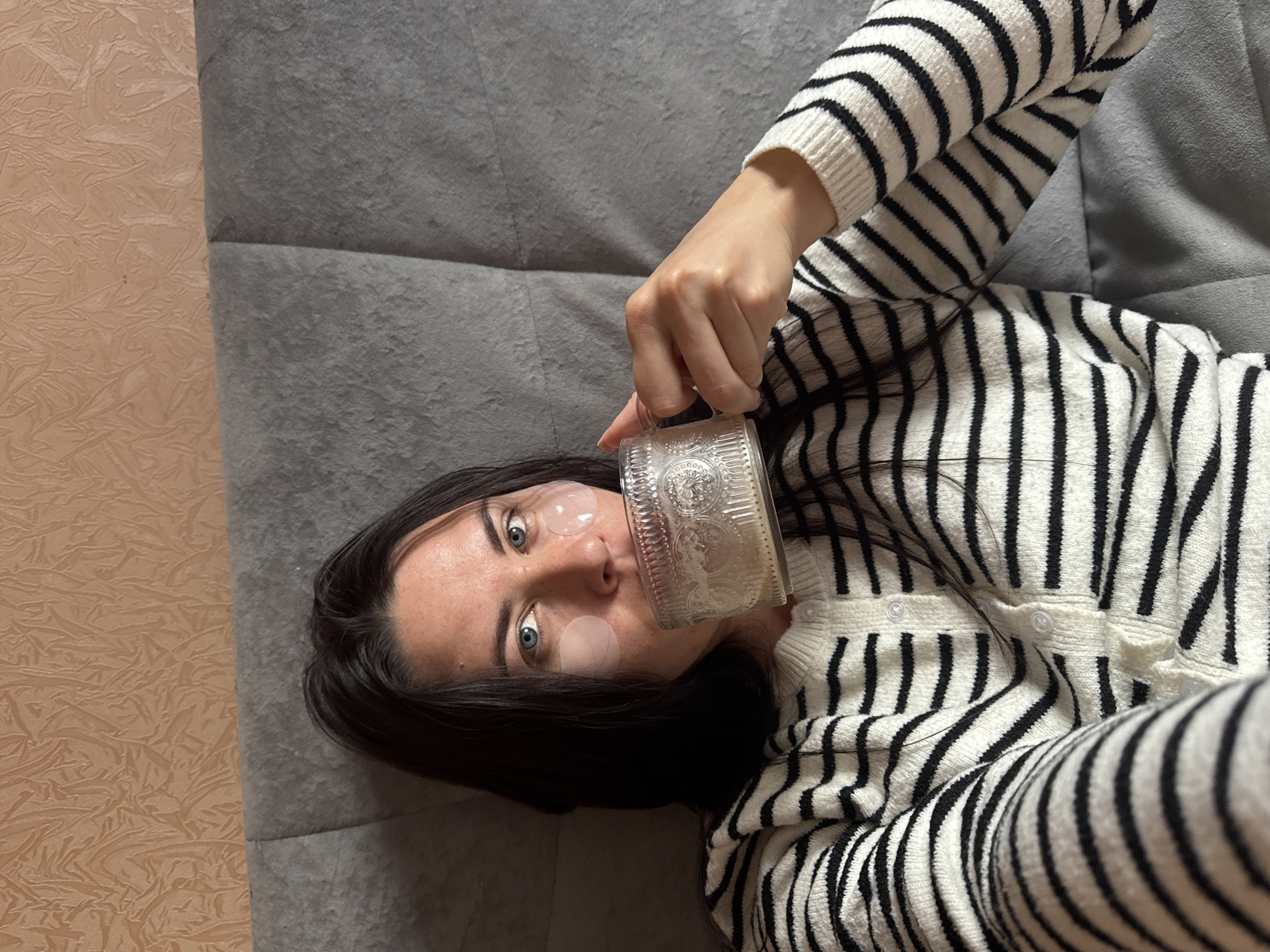 A woman with dark hair and blue eyes is lying down, holding a clear glass mug with both hands and drinking from it. She is wearing a white and black striped cardigan. Under her eyes, she has clear eye patches. She is positioned on a grey upholstered headboard, with a textured peach-colored wall behind it. The scene suggests a relaxed, personal moment within a home environment in the city of Chaikovsky, Russia.