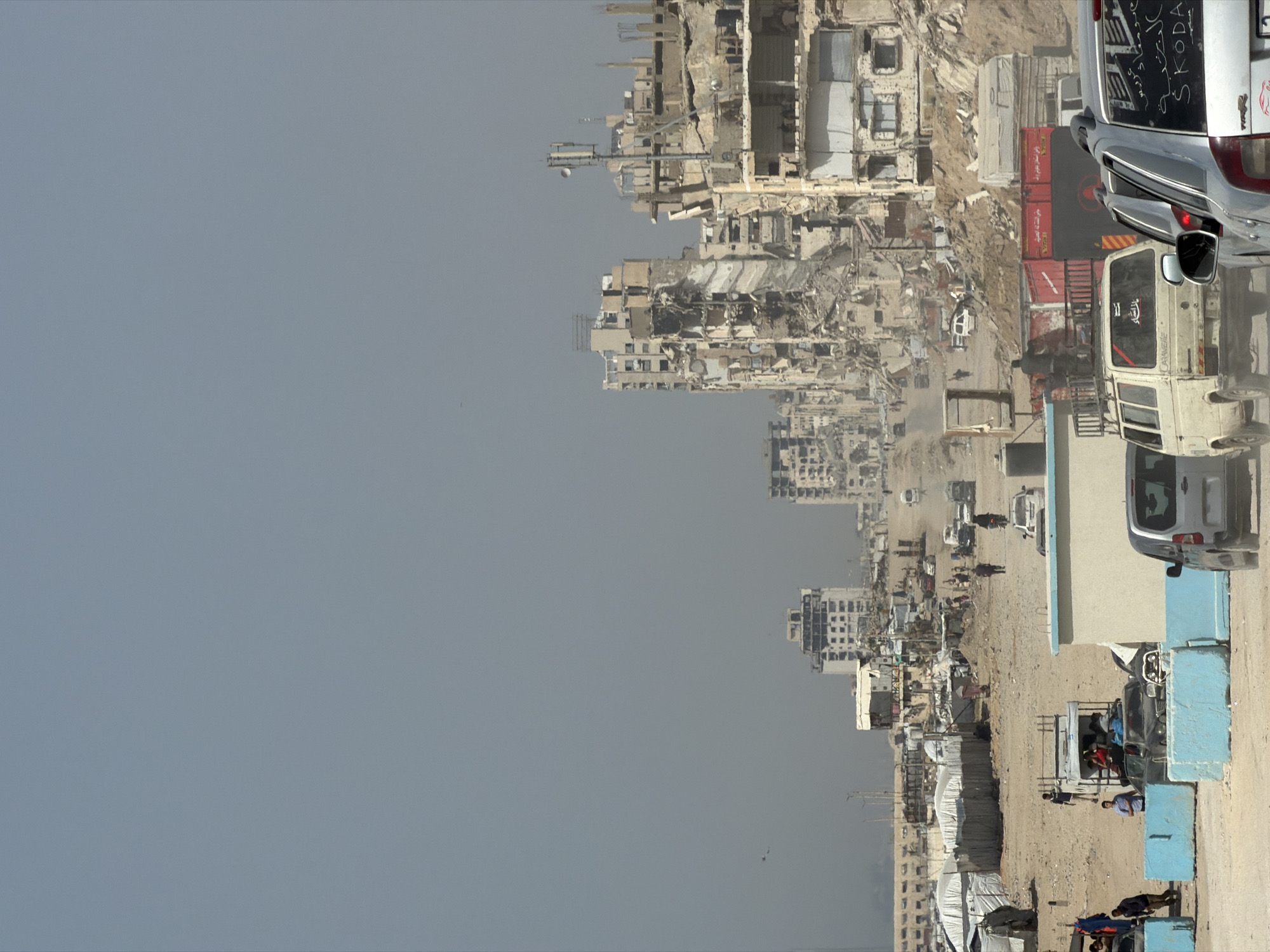 This image depicts a scene of destruction and displacement in an unknown city. On the left side of the frame, extensive damage to buildings is visible, suggesting the aftermath of a major destructive event, possibly an earthquake or conflict. The buildings are reduced to rubble and skeletal structures.
On the right side, a makeshift settlement or refugee camp is set up in a relatively open area. Numerous tents and temporary shelters are visible. People are seen engaging in various activities, possibly including resting, conversing, or tending to daily needs. Several vehicles are present, including cars, vans, and trucks, some of which appear to be involved in aid distribution or transportation. Some of the vehicles appear to be heavily laden. The ground is sandy and dusty, and the sky is overcast or hazy, creating a somber atmosphere.