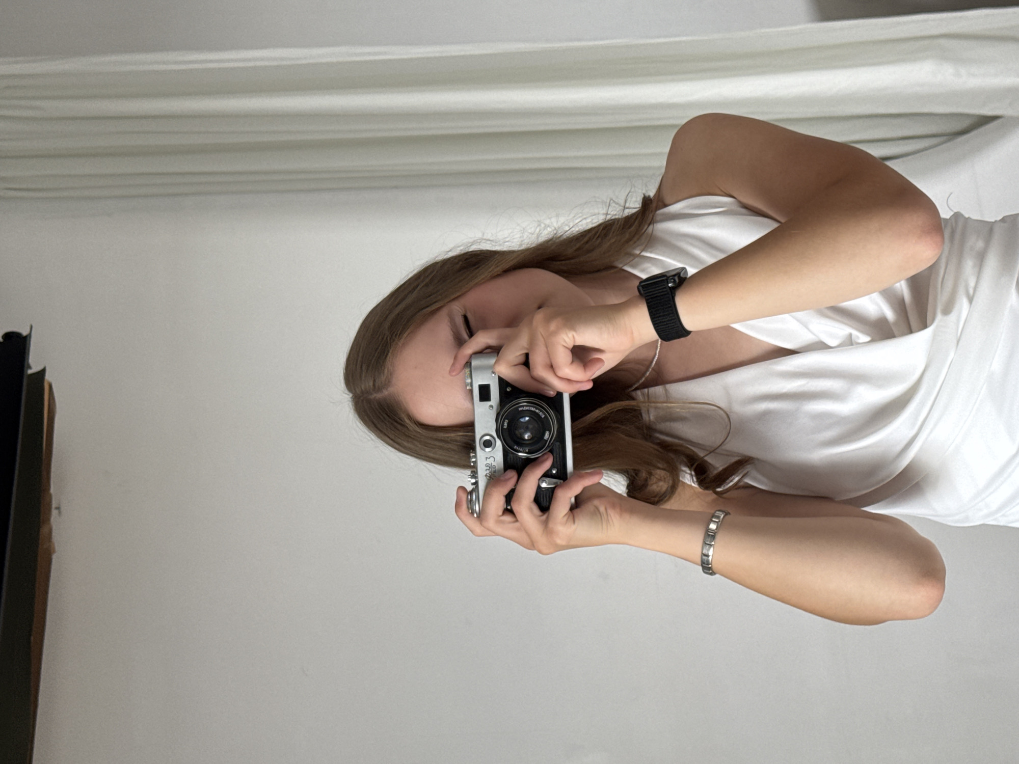 A young woman with light brown hair is holding a vintage camera to her face, looking through the viewfinder. She is wearing a white, draped dress and a black watch on her left wrist, as well as a silver bracelet on her left arm. The background is a plain white wall with a white curtain on the right side. The lighting is soft and diffused. The scene is indoors, likely in a studio or well-lit room, and there is no indication of the city of Chaikovsky, Russia, in the image itself.