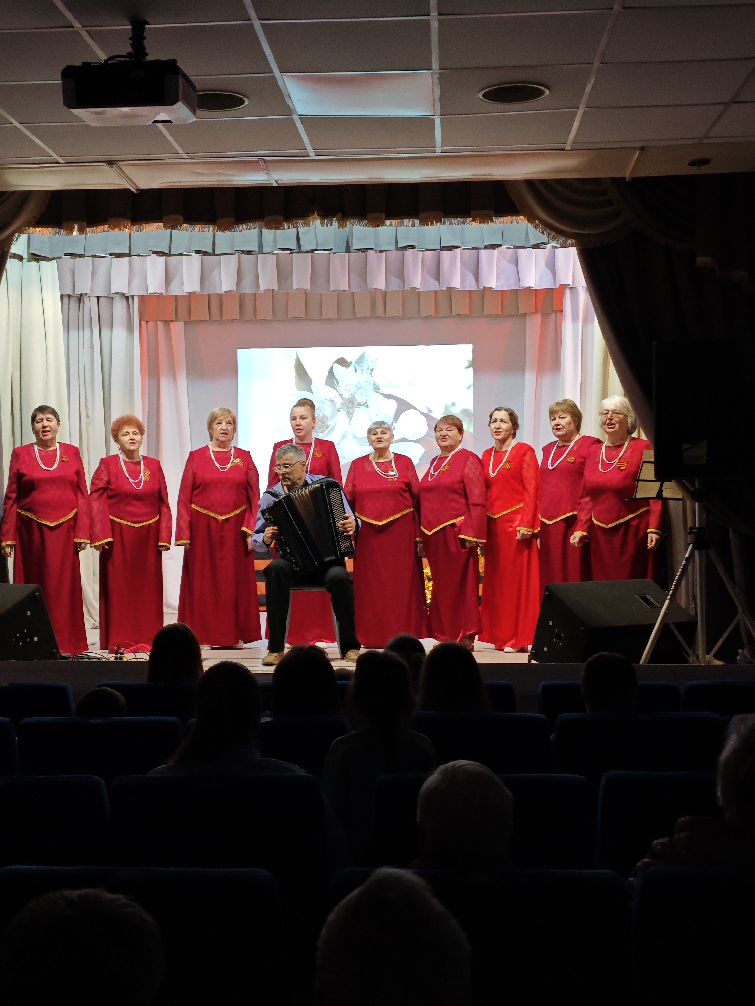 On a stage in Фоки, Russia, a choir of nine women in red dresses with gold trim and pearl necklaces sing while a man plays an accordion. The stage has a backdrop of white curtains and a screen displaying a floral image. Brown curtains hang at the top of the stage. The choir and musician are performing for an audience seated in blue chairs. A projector is visible on the ceiling.