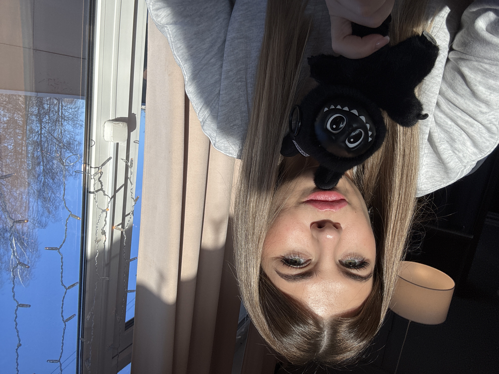 A young adult female is depicted indoors, positioned in front of a window. She has long, straight, light brown hair, green eyes, and is wearing a light grey long-sleeved sweatshirt. She holds a small, black plush toy with prominent white eyes and a wide, toothed smile. Sunlight streams from the right, illuminating her face and casting shadows. In the background, to the left, a light-colored drum-shaped pendant lamp hangs from a dark ceiling. To the right, through a window, a clear blue sky is visible, along with bare tree branches. Decorative string lights with small bulbs are affixed vertically to the interior side of the windowpane. The exact location within the city of Chaikovsky, Russia, is not identifiable from the provided visual evidence.
