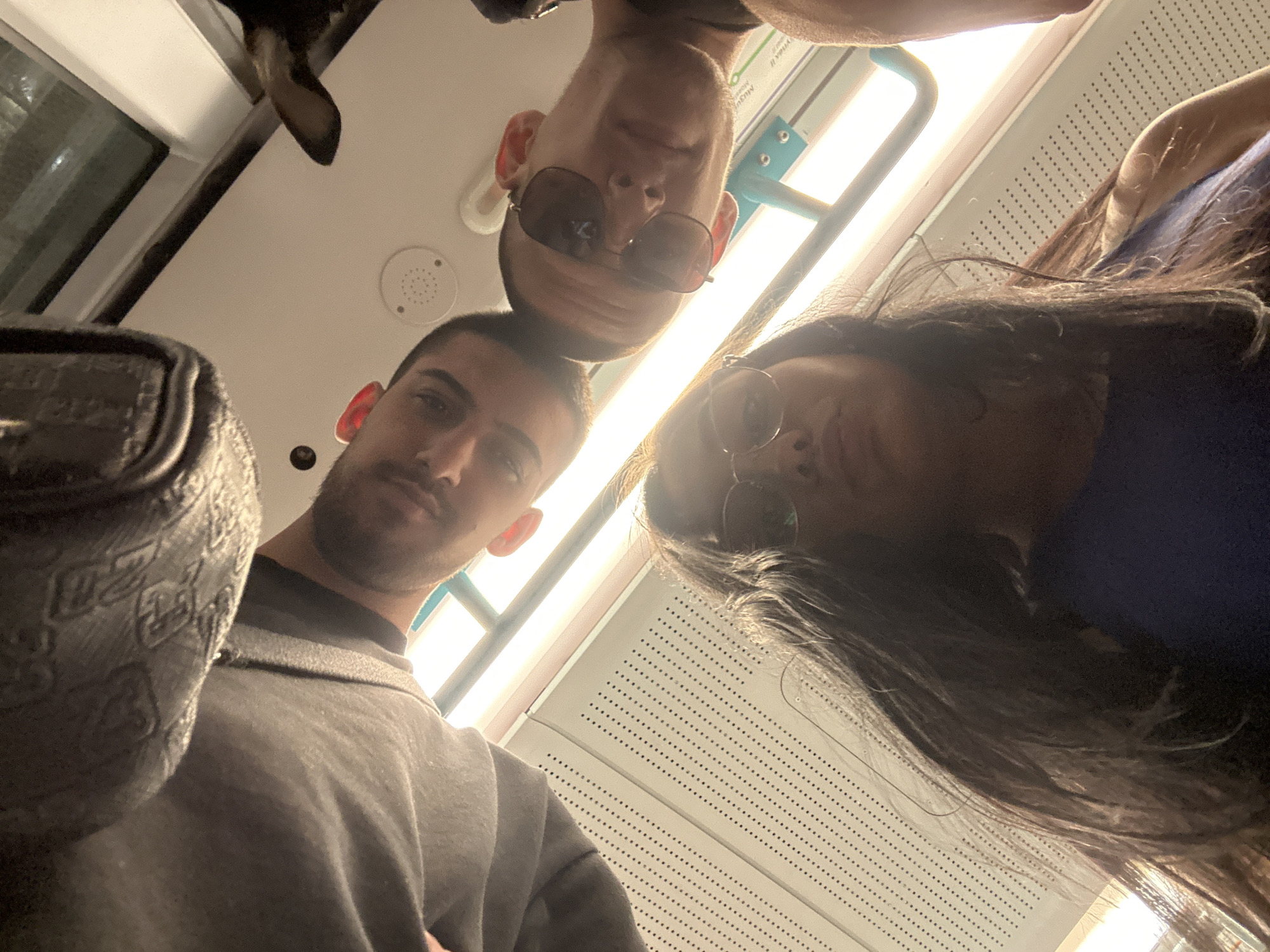 The image is a selfie taken from a low angle on a public transport vehicle, likely a train or bus. Three individuals are visible, two men and one woman, all looking down at the camera.
The man in the foreground on the right is wearing sunglasses and a grey t-shirt. He has short hair and his right ear is visible.
The man to his left, further back, is wearing a grey hoodie. His left ear is visible.
The woman in the foreground, at the bottom of the frame, has long dark hair and is wearing glasses and a blue top.
The background shows the interior of the vehicle, with fluorescent lighting, a perforated panel on the wall, and a metal handrail. There is also a glimpse of a dog's ear in the upper right corner. The overall lighting is bright and somewhat harsh. The image is oriented such that the heads of the individuals are pointing downwards.