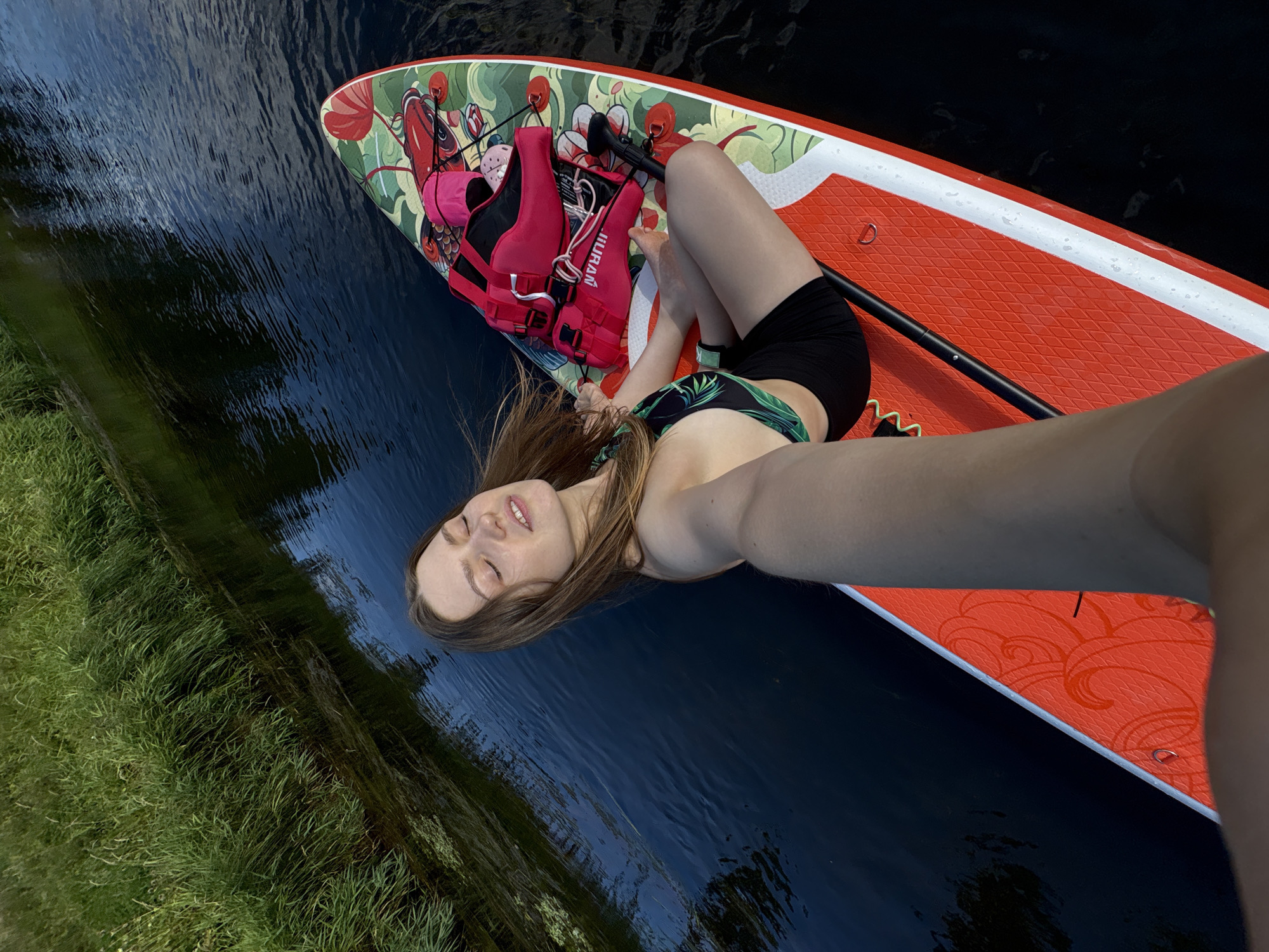 A young woman is lying on a paddleboard in dark blue water, with green grass on the left side of the frame. The paddleboard is orange and has a decorative design of a koi fish and flowers in red, green, and white. The woman is wearing a black bikini with a tropical leaf pattern and a black watch. A pink life vest is also visible on the paddleboard. She is looking up at the camera with her eyes closed and a slight smile on her face, suggesting a relaxed and peaceful mood. Her long brown hair is spread out on the paddleboard. The scene appears to be set during the daytime, with natural light illuminating the water and the paddleboard. The presence of grass and water suggests an outdoor recreational setting, possibly a lake or calm river. The visible text on the life vest reads "HILIRAN." The overall impression is one of leisure and enjoyment in a natural environment.