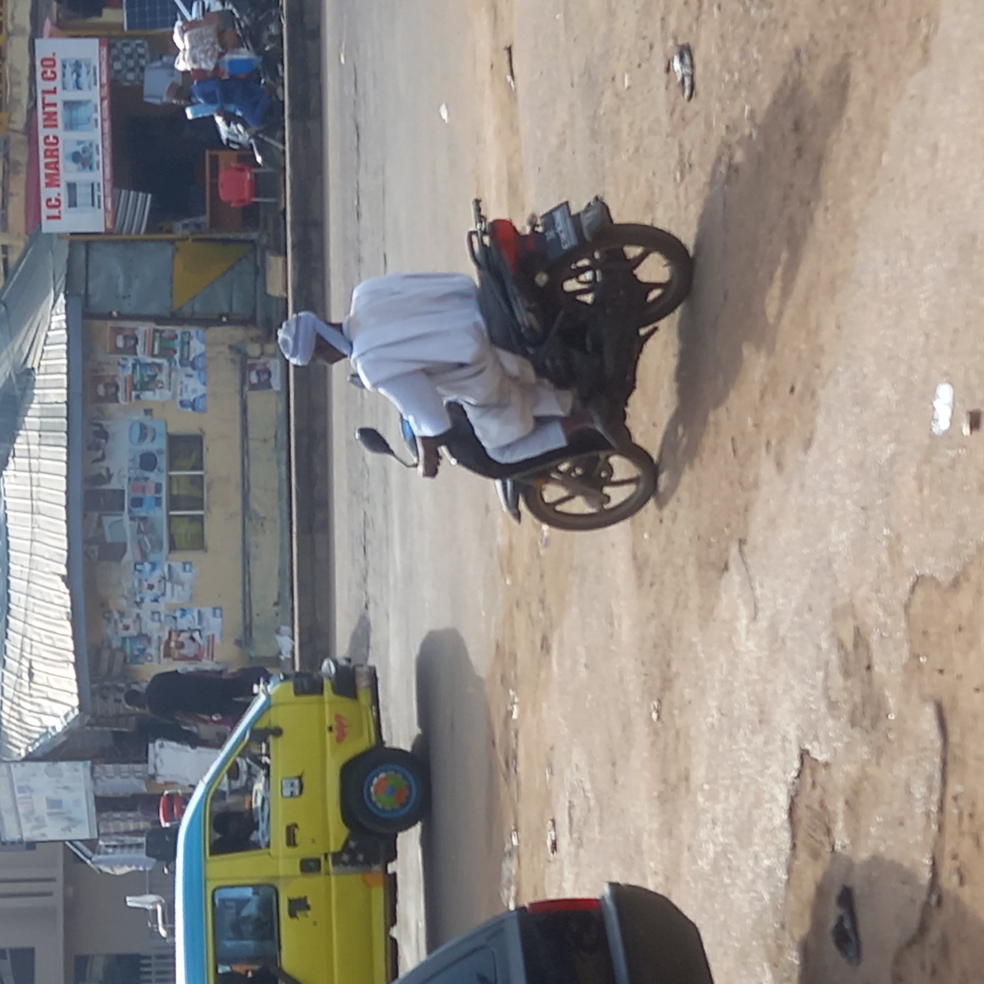 A man wearing a white robe and cap sits on a black motorcycle in Dadi Riba, Nigeria. The motorcycle is positioned on a dirt road. To the left, a yellow and blue bus is visible. A store with the sign "LC. MARC INT'L CO." is also on the left, with posters affixed to the wall. A person in dark clothing walks past the bus. A portion of a dark car is visible in the lower foreground.