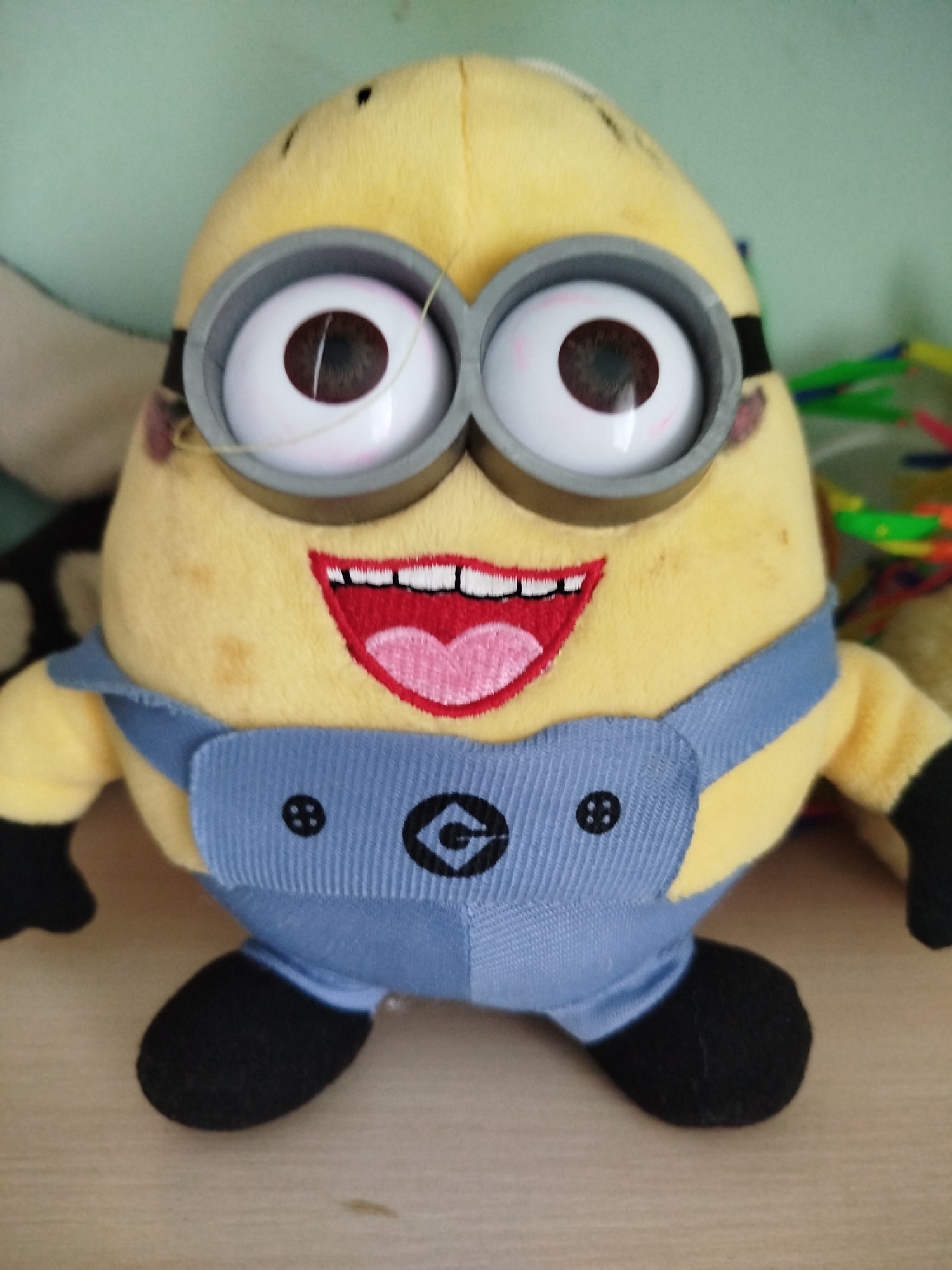 A stuffed Minion toy is positioned centrally in the frame, set against a light blue wall and a light brown wooden surface. The toy is yellow with large, round goggles, a wide smiling mouth, and is wearing blue overalls with a black button and a black Minion logo on the front. The background shows out-of-focus colorful objects, possibly toys or art supplies, on the right. The lighting is even, with no harsh shadows. The image does not depict any specific location in Крывасельскі сельскі Савет, Belarus.