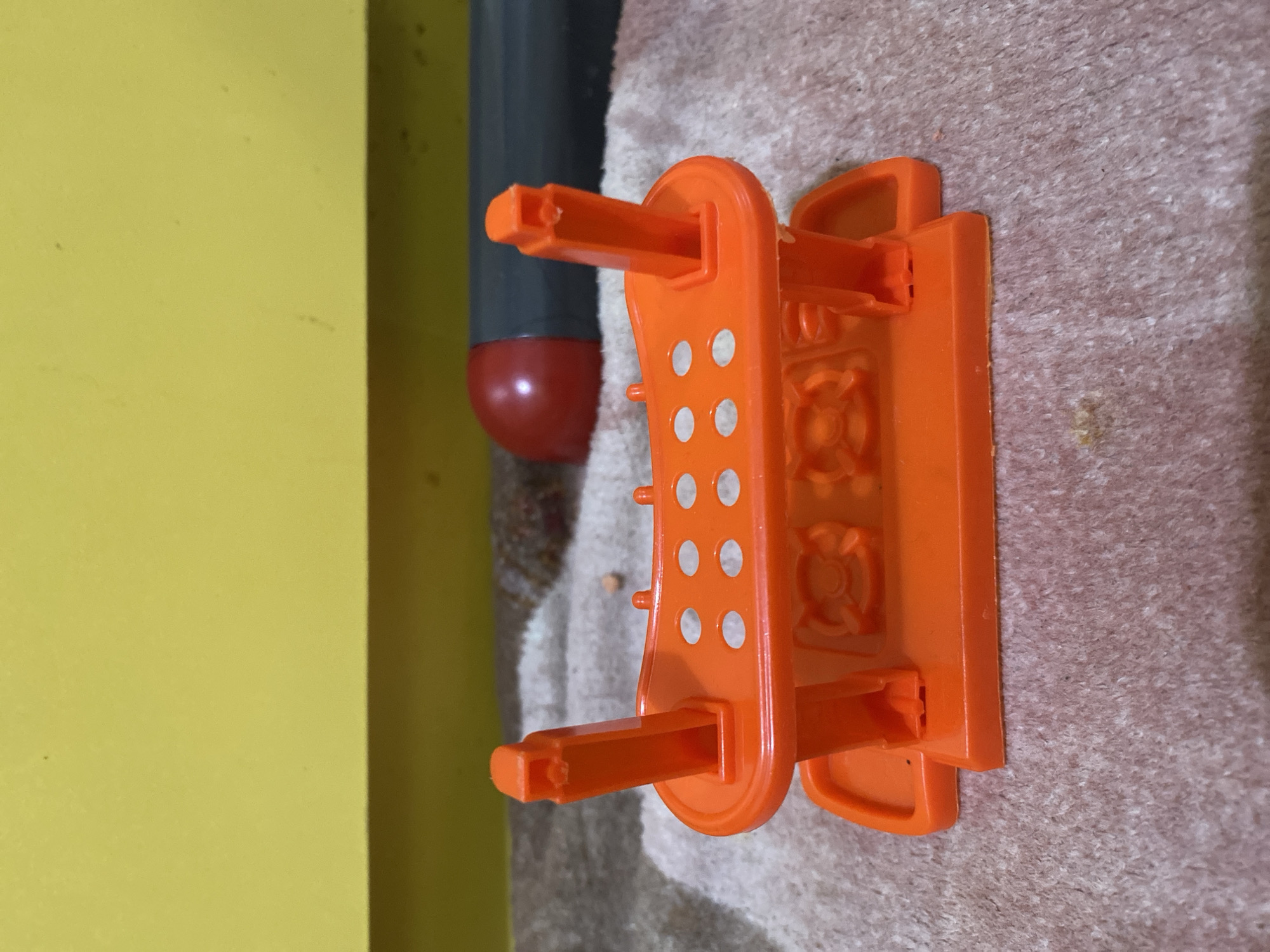 An orange plastic laboratory rack is situated on a textured, pinkish-brown surface. The rack has six circular holes on its upper platform, designed to hold test tubes. Two vertical supports rise from each side of the base, and two similar supports are at the front. The base also features two circular indentations with a four-lobed design. In the background, a yellow surface is visible above a dark gray cylinder with a red tip. The scene is indoors, likely in Lambaro Kueh, Indonesia, though no specific urban features are discernible. There are no people present.