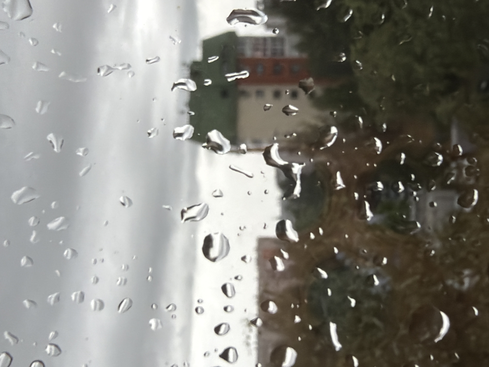 The image depicts a view through a window covered in raindrops. The background is blurred, showing buildings and trees, suggesting an urban or suburban environment. The sky appears overcast and gray, consistent with rainy weather. The focal point of the image is the distorted view of the outside world as seen through the water droplets on the glass. There are no people or specific identifiable actions captured in the scene. Without additional information, it is not possible to determine the exact location within Surgut, Russia.