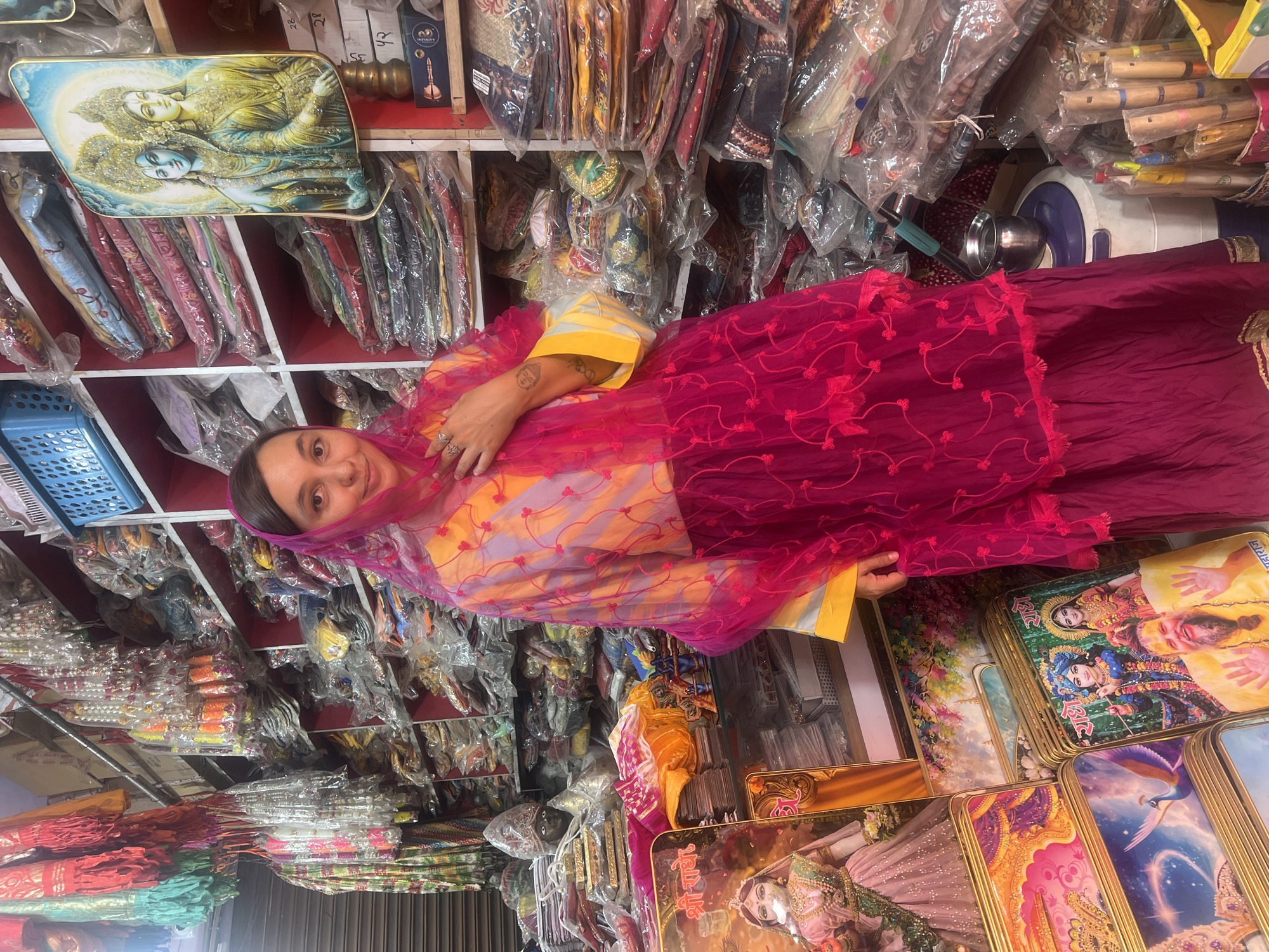 A woman wearing a traditional Indian dress and head covering stands in a shop filled with religious merchandise in Mathura, India. The shop has shelves stocked with packaged fabrics and other items. The woman is fair-skinned, and the translucent pink head covering partly obscures her face. Her dress is a combination of yellow, lavender, and dark pink. Her left arm is extended, showing tattoos. The foreground is dominated by a stack of framed religious images. A framed depiction of Hindu deities is visible on the upper left.