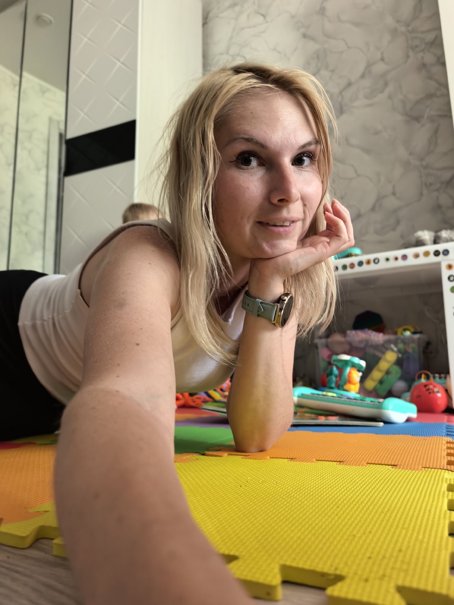 A woman is lying on a brightly colored foam mat, with her head resting on her hand. She has blonde hair and is wearing a white tank top. In the background, there is a shelf filled with toys, and a child is partially visible. The environment appears to be a playroom or nursery, likely in a residential setting within the city of Sterlitamak, Russia.
