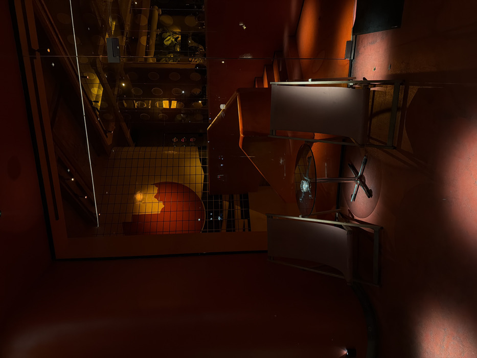 A surreal, orange-toned interior scene is depicted. A structure with circular, patterned surfaces is visible above. Below, a wireframe cage encloses a red and yellow object. A series of orange slides lead towards two tables, each with a bicycle wheel attached. The scene is characterized by strong shadows and an unconventional perspective.