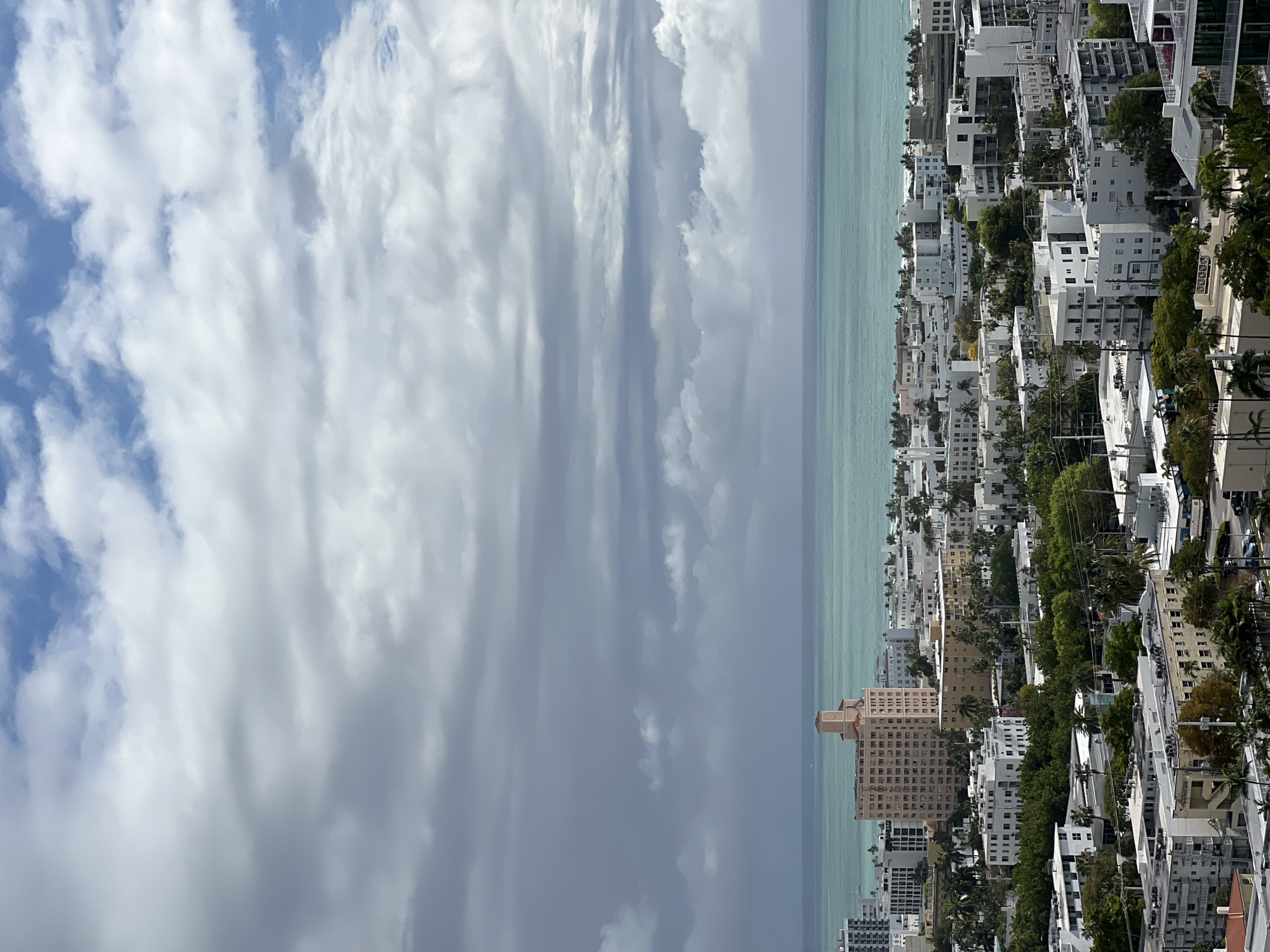 The image depicts an aerial view of Miami Beach, Florida, USA. The frame is split into three primary horizontal sections. The leftmost section is the sky, filled with predominantly gray and white clouds under a blue sky. The middle section is the ocean, rendered in a light turquoise color. The rightmost section shows the cityscape of Miami Beach. The buildings are primarily white. Green trees are interspersed among the buildings. The horizon line is the boundary between the ocean and the sky.