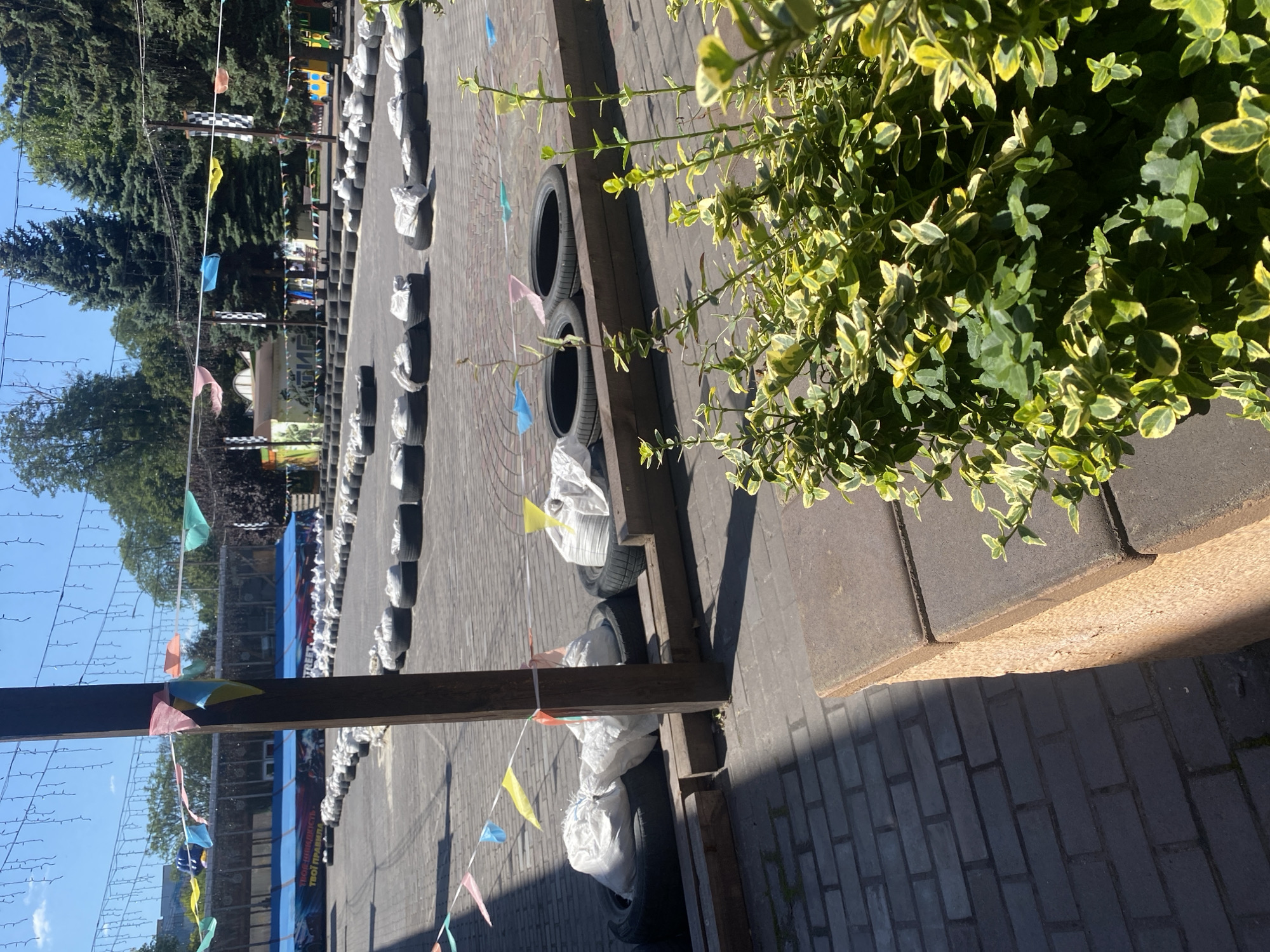 The image depicts an outdoor go-kart track in the city of Kamianske, Ukraine, on a clear, sunny day. The track is lined with tires, some of which are filled with white bags, serving as barriers. Colorful triangular flags are strung across the track and on poles, indicating a festive or event atmosphere. In the background, there is a building with a blue awning and signage, suggesting it is a commercial establishment, possibly related to the go-karting activity. Trees line the perimeter of the area, and the ground is paved with brick-like material. In the foreground, lush green and yellow foliage is visible, partially obscuring the view of the track. There are no people visible in the image.