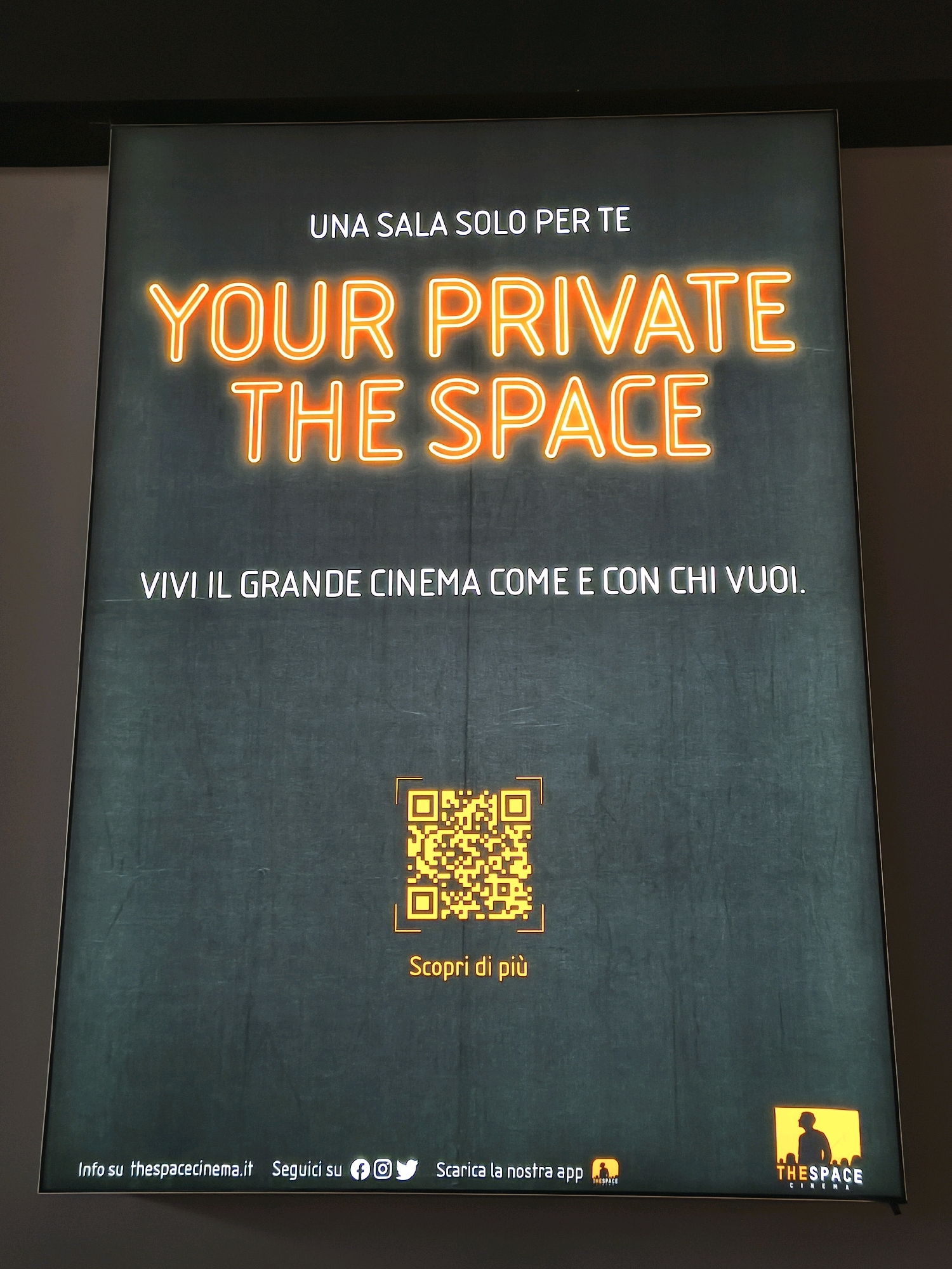 The image shows a large advertisement on a dark, textured background, likely a screen or a billboard. The advertisement features bright orange neon text that reads "YOUR PRIVATE THE SPACE" in large, prominent letters. Above this, smaller white text states "UNA SALA SOLO PER TE". Below the neon text, another line of white text says "VIVI IL GRANDE CINEMA COME E CON CHI VUOI."
Centered in the lower half of the advertisement is a yellow QR code. Below the QR code, in orange text, is "Scopri di più".
At the very bottom of the advertisement, a white text bar contains information: "Info su thespacecinema.it Segiuici su" followed by social media icons for Facebook, Instagram, and Twitter. To the right of these icons, white text reads "Scarica la nostra app", with a small orange logo featuring a silhouette of a person. In the bottom right corner, a larger logo with the text "THESPACE CINEMA" in orange and white is displayed, also with a silhouette of a person within an orange box.
The lighting suggests an indoor setting, possibly a cinema lobby or a public space where such advertisements are displayed. The image does not contain any specific visual cues that would allow for the identification of its location in Chiugiana, Italy.