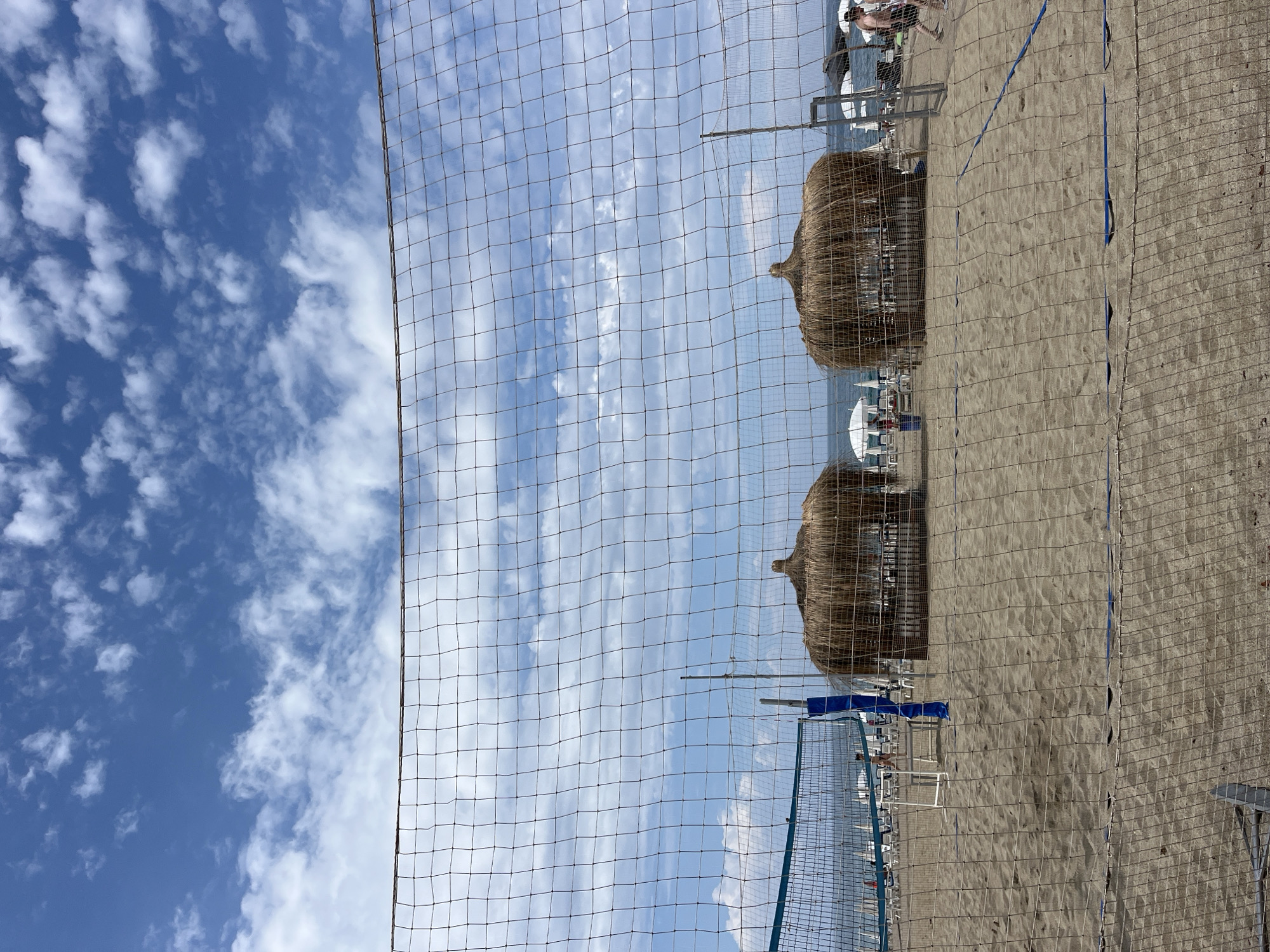 The image shows a beach scene in Alanya, Turkey, viewed through a volleyball net. The sand is light brown, and in the background are two straw beach umbrellas providing shade to beachgoers. Beach chairs and tables are visible under the umbrellas, along with some people. Beyond the beach, the blue sky is dotted with white clouds. The image is shot at an angle, tilting the horizon line to the right.