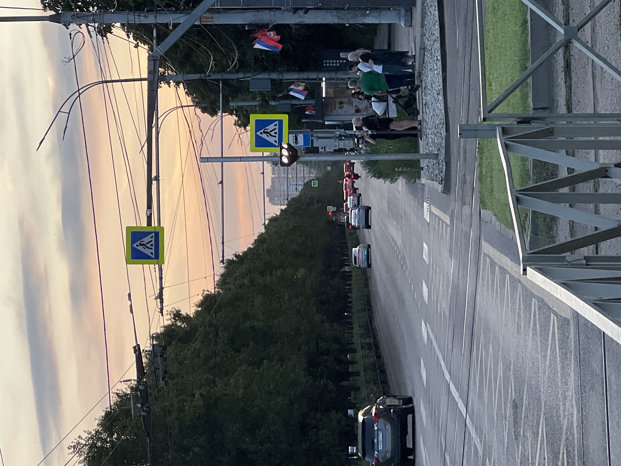 The image shows a street scene in Krasnodar, Russia, at dusk. The sky is filled with hues of orange, pink, and purple from the setting sun. A wide asphalt road with multiple lanes is visible, marked with white lines. There are tram tracks embedded in the road. On the left side of the road, a dense row of tall, dark green trees lines the street, with some vegetation in front of them. On the right side of the road, there is a sidewalk with a group of people standing. Among them are two women in black, one pushing a baby stroller, a man in a green t-shirt, a man in a white t-shirt, and a woman in a dark dress. Behind them is a building with a shop sign that appears to say "ВКУС" (Vkus). Further down the street, several cars are driving, including a dark-colored SUV in the foreground on the left. Traffic lights and pedestrian crossing signs are visible, including a pedestrian crossing sign with a blue background and a white silhouette of a person walking. Several power lines and poles crisscross the sky above the street. Russian flags are hanging from a pole on the right side of the image. The overall impression is a typical urban street in the evening.