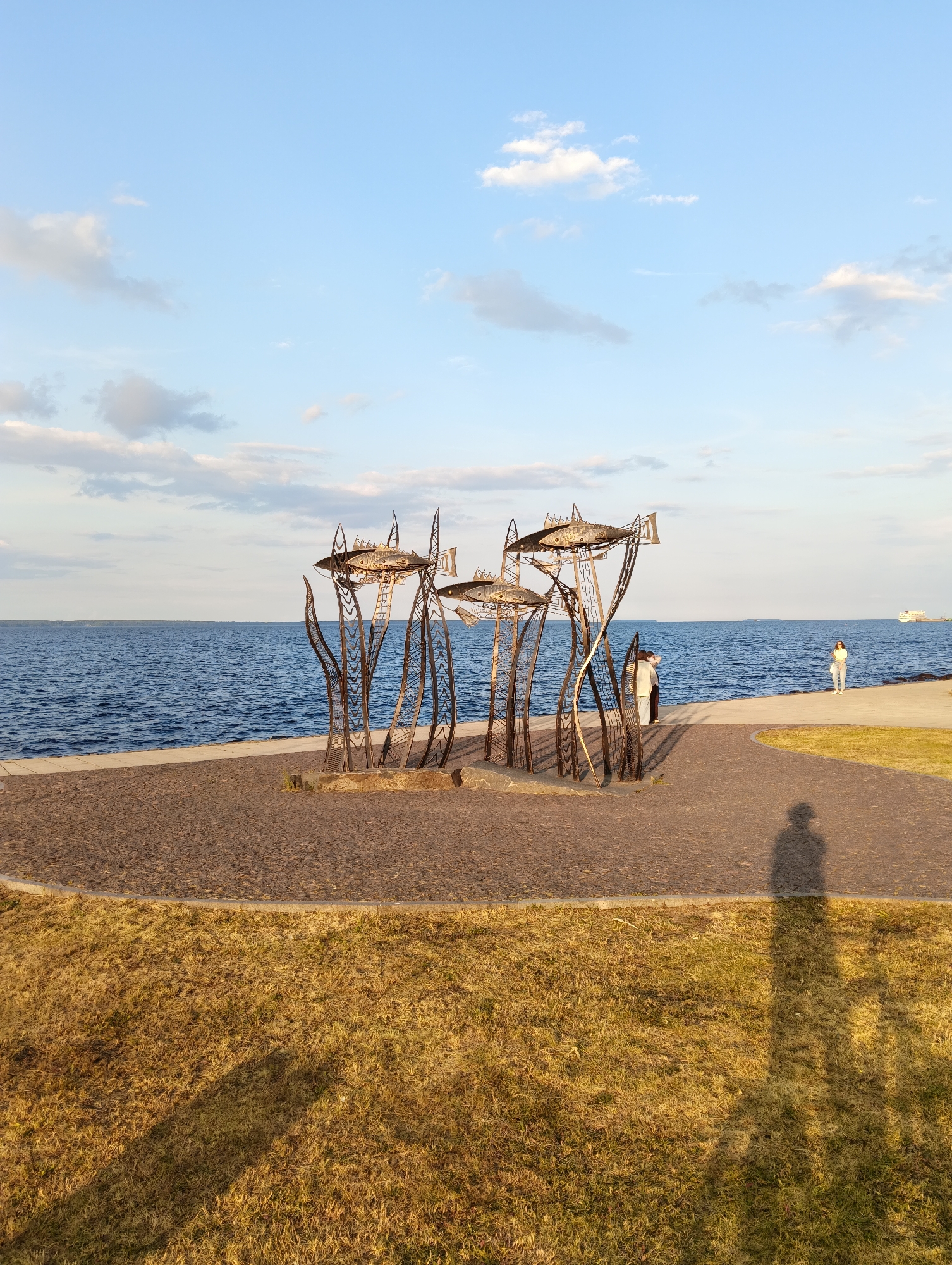 This image depicts a waterfront scene at Onega Lake in Petrozavodsk, Russia, during what appears to be late afternoon or early evening, given the lighting. In the foreground, a patch of dry, golden grass is visible, casting a long shadow of the photographer. Adjacent to the grass is a walkway made of dark gravel and stone, leading towards the water.
Dominating the middle ground is an abstract metal sculpture resembling stylized reeds or stalks topped with metallic fish-like figures. These sculptures are positioned on a circular area covered in gravel.
Further into the scene, the vast blue expanse of Onega Lake stretches to the horizon, with a faint treeline visible in the far distance. A paved promenade runs along the edge of the lake, separating it from the gravel area with the sculpture.
On the promenade, two individuals are standing near the sculpture, facing away from the camera. Further along the promenade, a lone woman in light-colored clothing is walking away from the sculpture and towards the right edge of the frame. In the distance, on the lake, a white ship is visible. The sky above is a clear, pale blue, with scattered white and grey clouds.