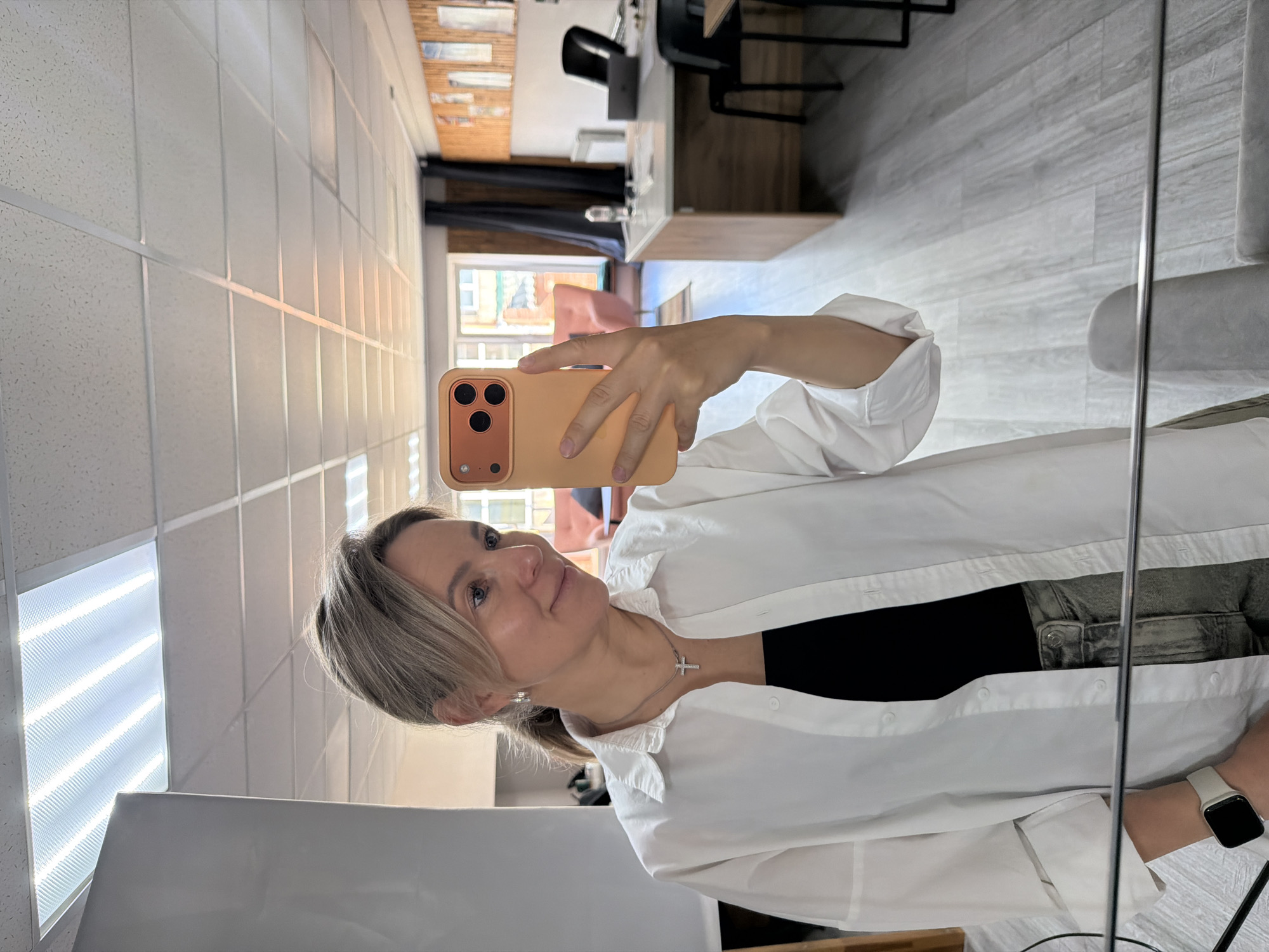 A woman is taking a selfie in a room with a window. She is wearing a white shirt, black top, jeans, and a silver cross necklace. She is holding a peach-colored smartphone with three camera lenses. Her hair is blonde and pulled back in a ponytail. She is wearing a smartwatch on her left wrist. The background of the image includes a ceiling with fluorescent lights, a gray floor, and a room with furniture. The location is implied to be in the city of Chaikovsky, Russia, but this is not explicitly visible in the image.