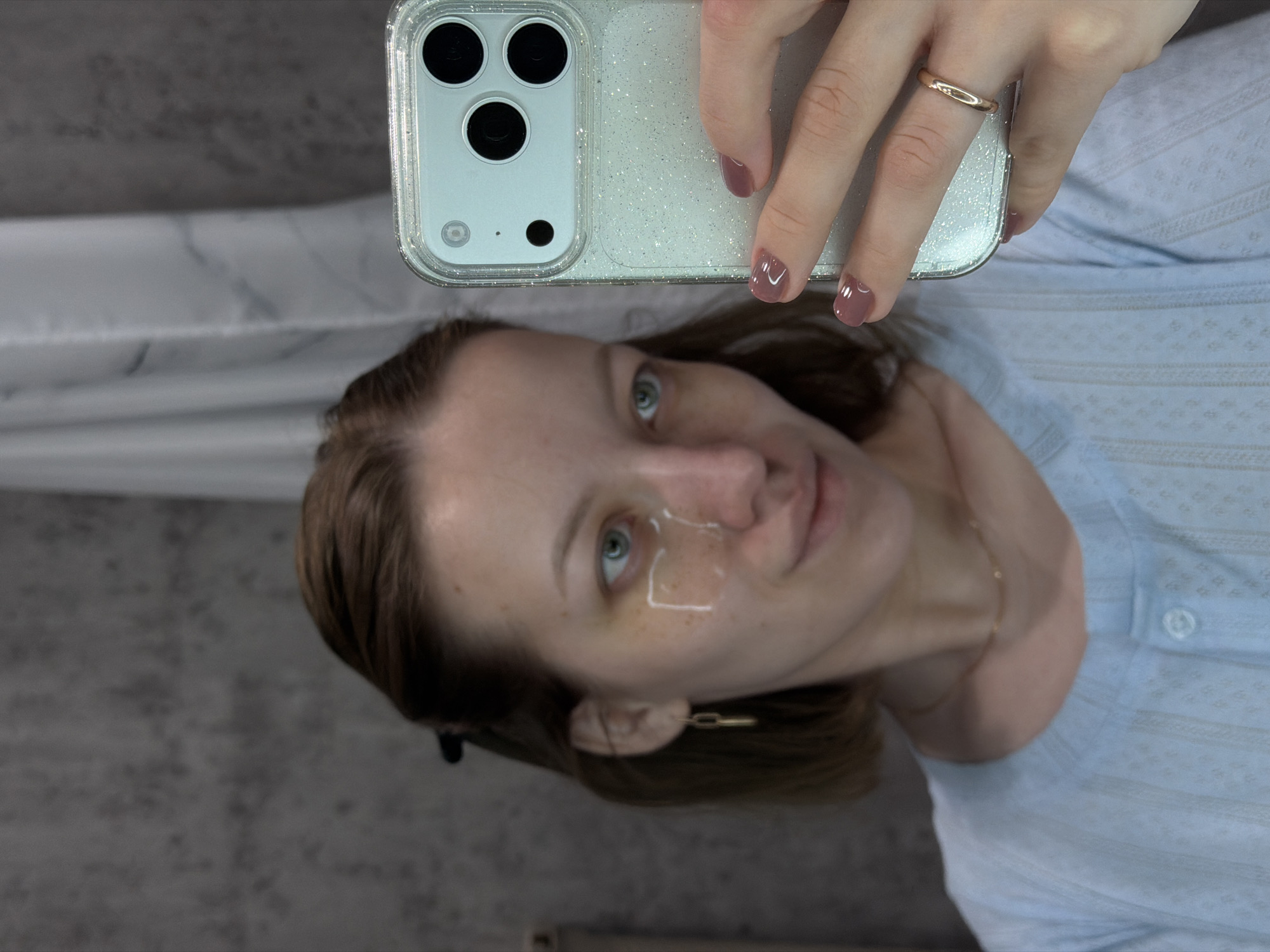 A woman takes a selfie with a phone in a clear, glittery case. She is wearing a light blue shirt. Her nails are painted a mauve color. She wears a gold ring and a gold chain earring. A clear patch is visible on her face, near her eye. The background appears to be a gray textured wall or surface.
There is no indication of the location as Reshetnikovo, Russia.