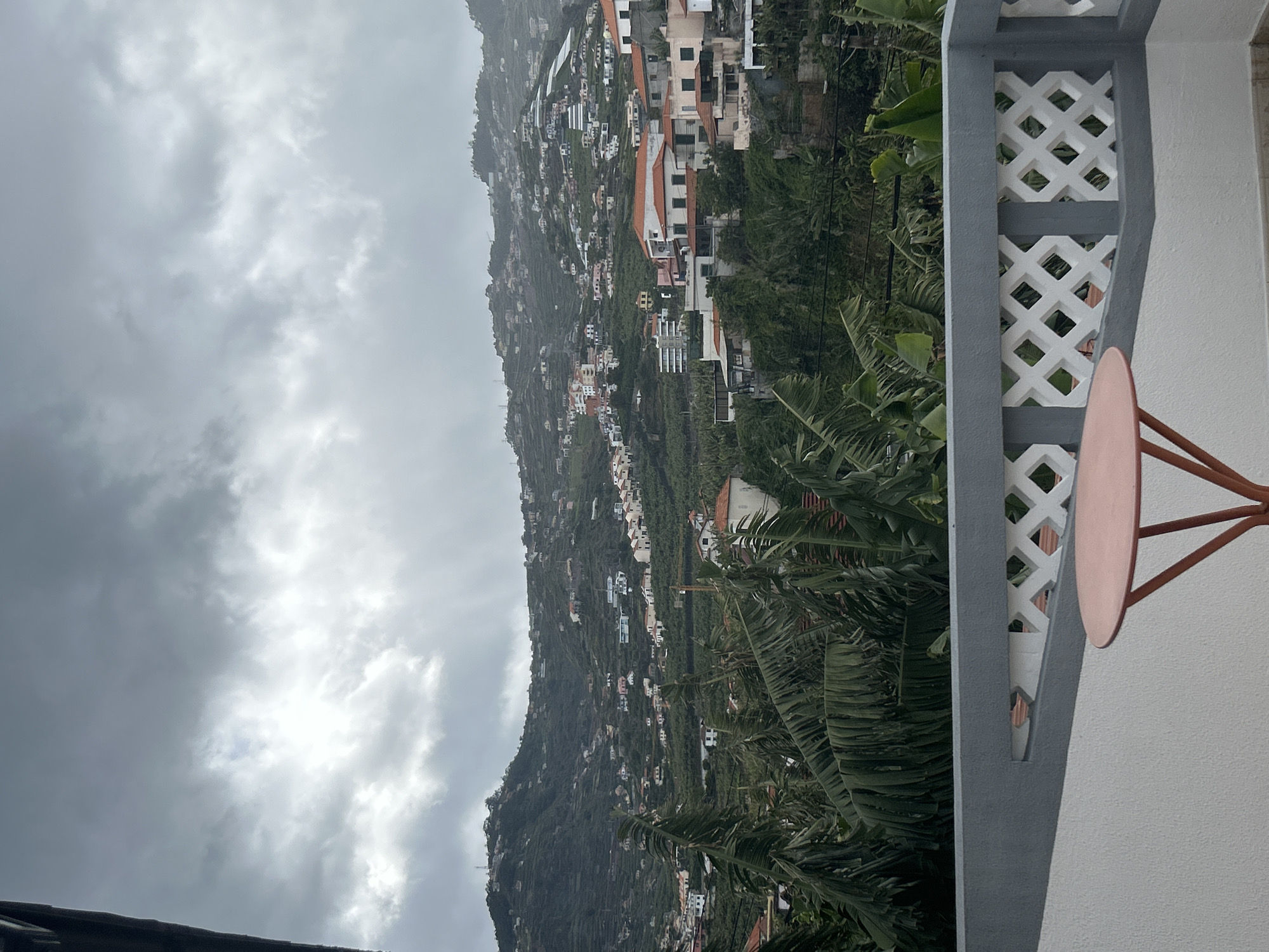 The image depicts a view from a balcony in Câmara de Lobos, Portugal, overlooking a hillside densely populated with houses. The hillside is covered in lush green vegetation, with banana trees and other greenery visible. The sky is overcast with dark, heavy clouds, suggesting a gloomy or rainy day.
In the foreground, there's a section of a white balcony with a grey lattice design and a small, round table with a peach-colored top and orange legs. The balcony serves as a partial frame, adding depth to the scene.
The houses on the hillside vary in size and color, with many featuring red tile roofs. They are clustered together and spread across the slope, indicating a built-up residential area. The image captures a sense of elevation and a sweeping view of the town clinging to the hillside.