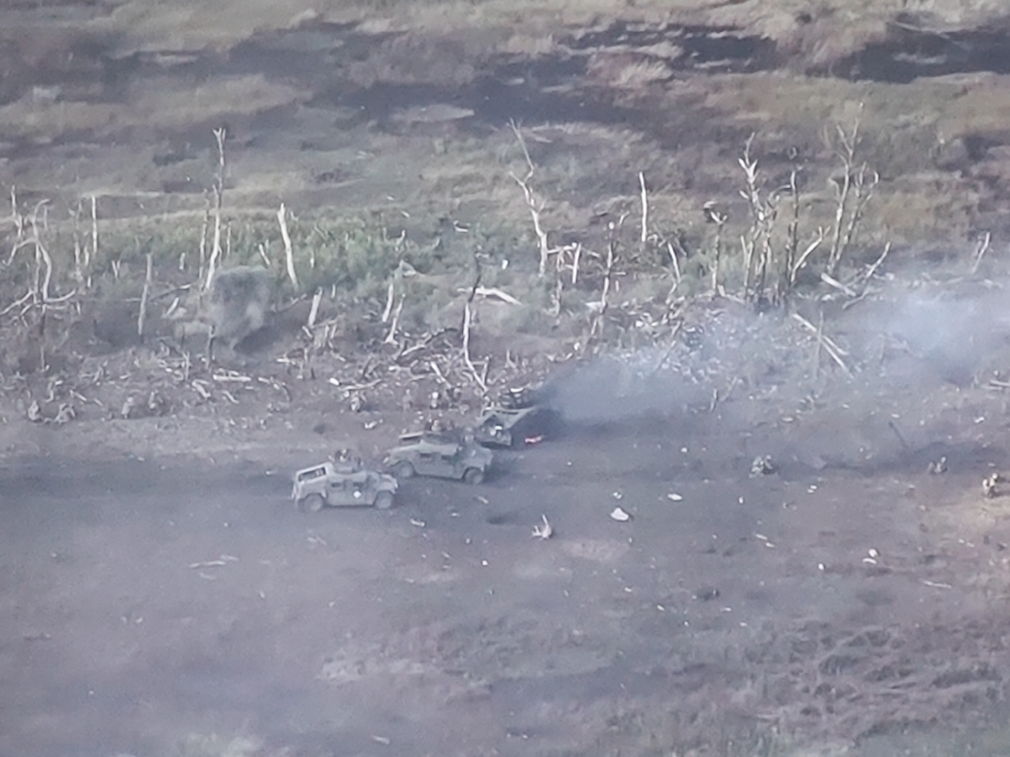 The image depicts an aerial view of a scene with military vehicles and smoke in a rural or overgrown area. There are at least three armored vehicles, two of which appear to be in operational condition, with one possibly being a humvee. Another vehicle, to the right, is emitting a significant amount of smoke, suggesting it is damaged or on fire. The ground is uneven and covered with debris, including broken branches and scattered white objects, possibly shell casings or small stones. In the background, there is sparse vegetation, including dead trees or branches, and patchy ground cover. The overall impression is of a recent engagement or a military operation in a somewhat desolate environment. The provided information about the location in Molenbeek-Saint-Jean - Sint-Jans-Molenbeek, Belgium, is not visually verifiable from the image content.